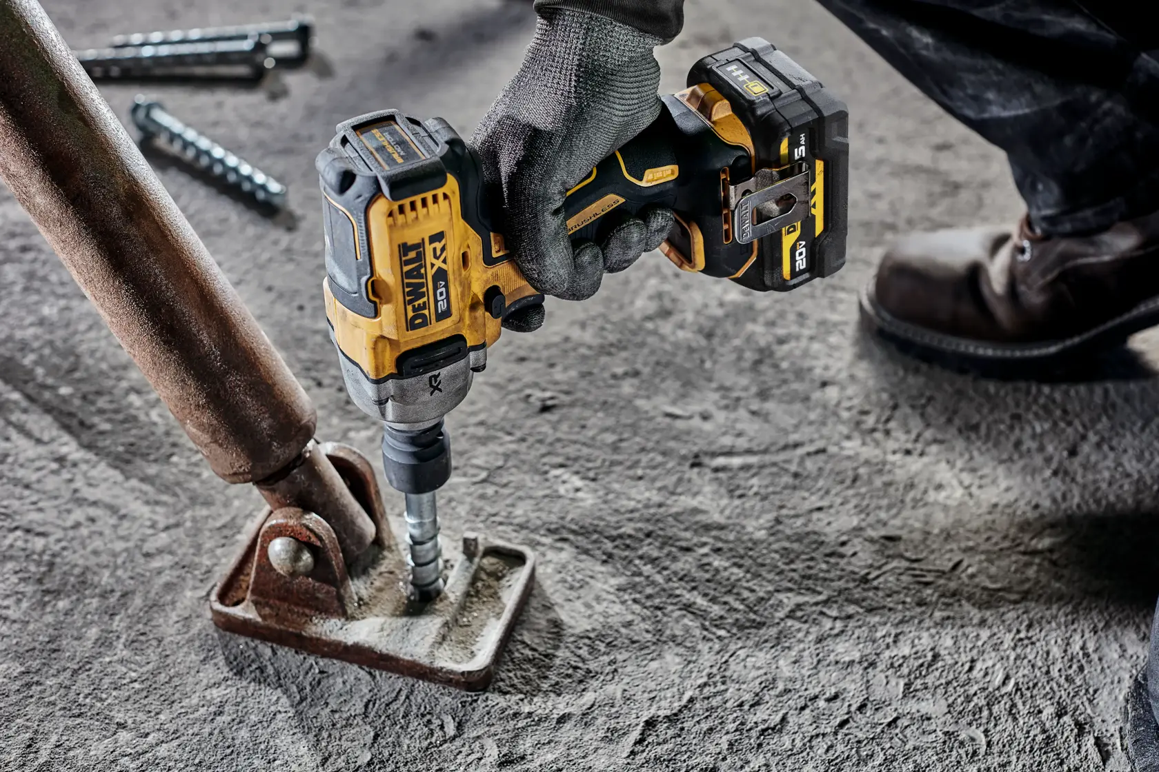 A person wearing gloves uses a DEWALT cordless impact wrench to fasten a bolt into a metal base plate on a rough concrete surface.