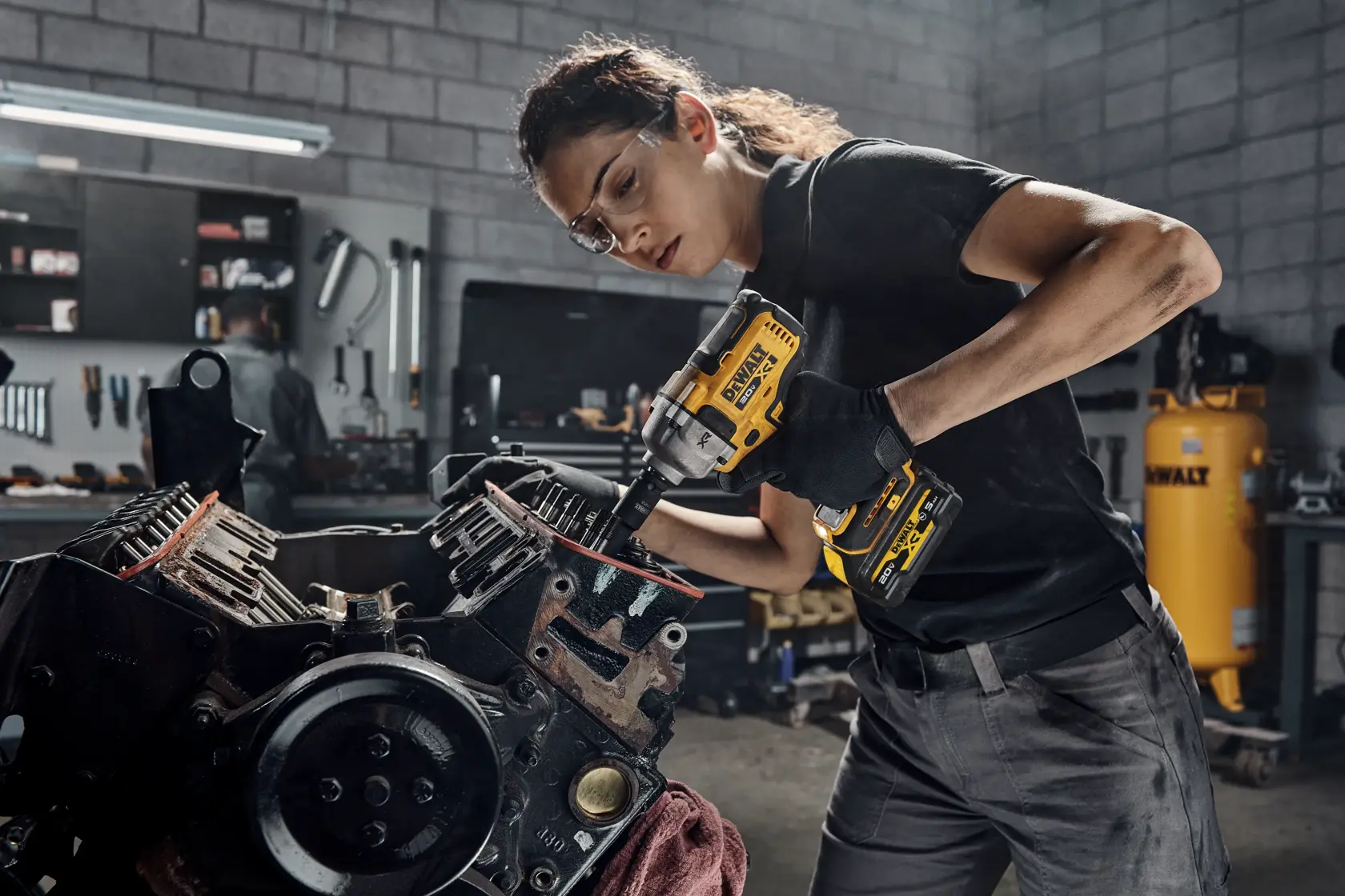 A person using a DEWALT cordless impact wrench to work on an engine in a workshop. The individual’s face is blurred for privacy. The workspace includes tools, shelves, and a DEWALT air compressor in the background.
