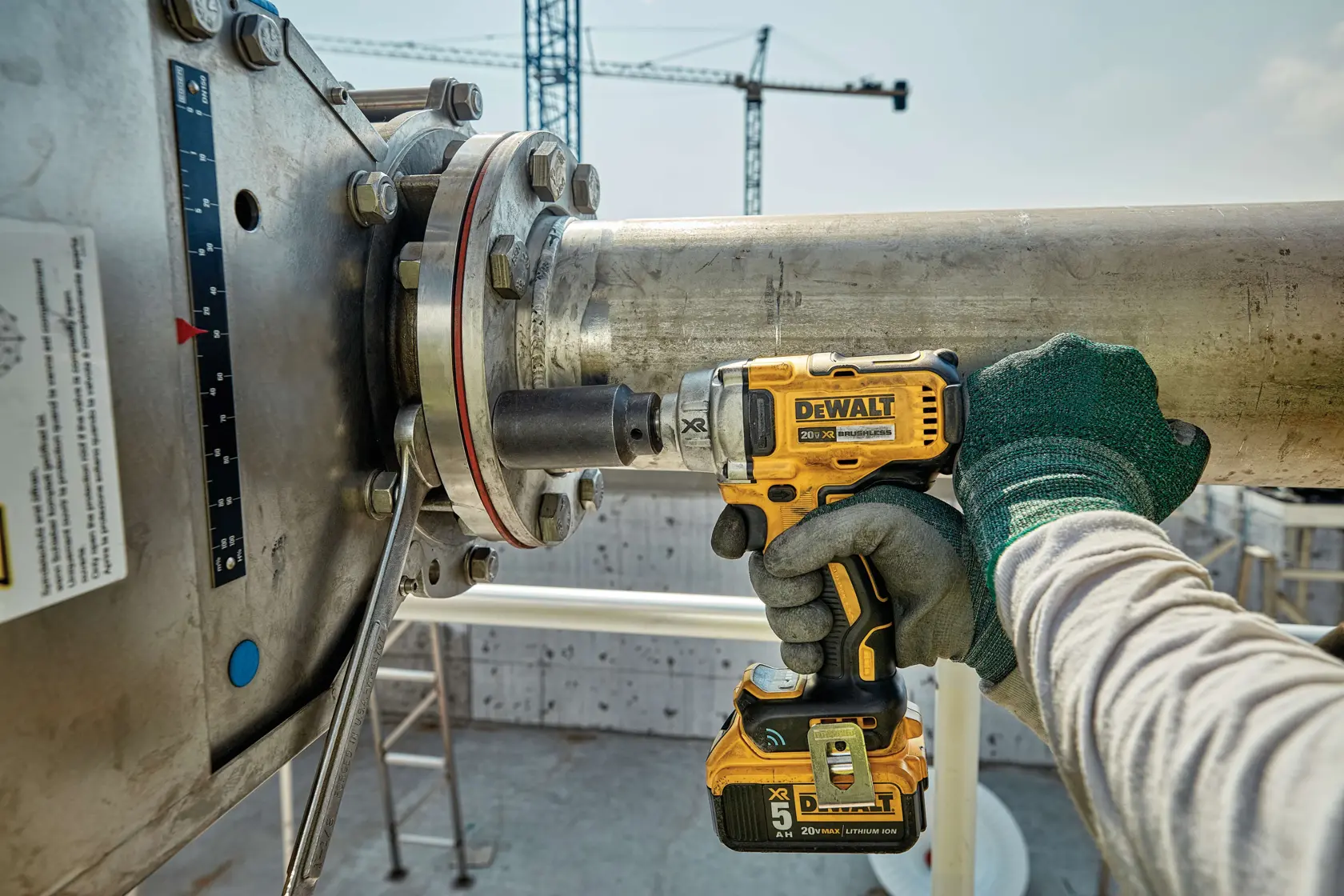 A worker uses a DEWALT cordless impact wrench to fasten or loosen bolts on a large industrial metal pipe outdoors. The worker is wearing green protective gloves and a long-sleeve shirt.