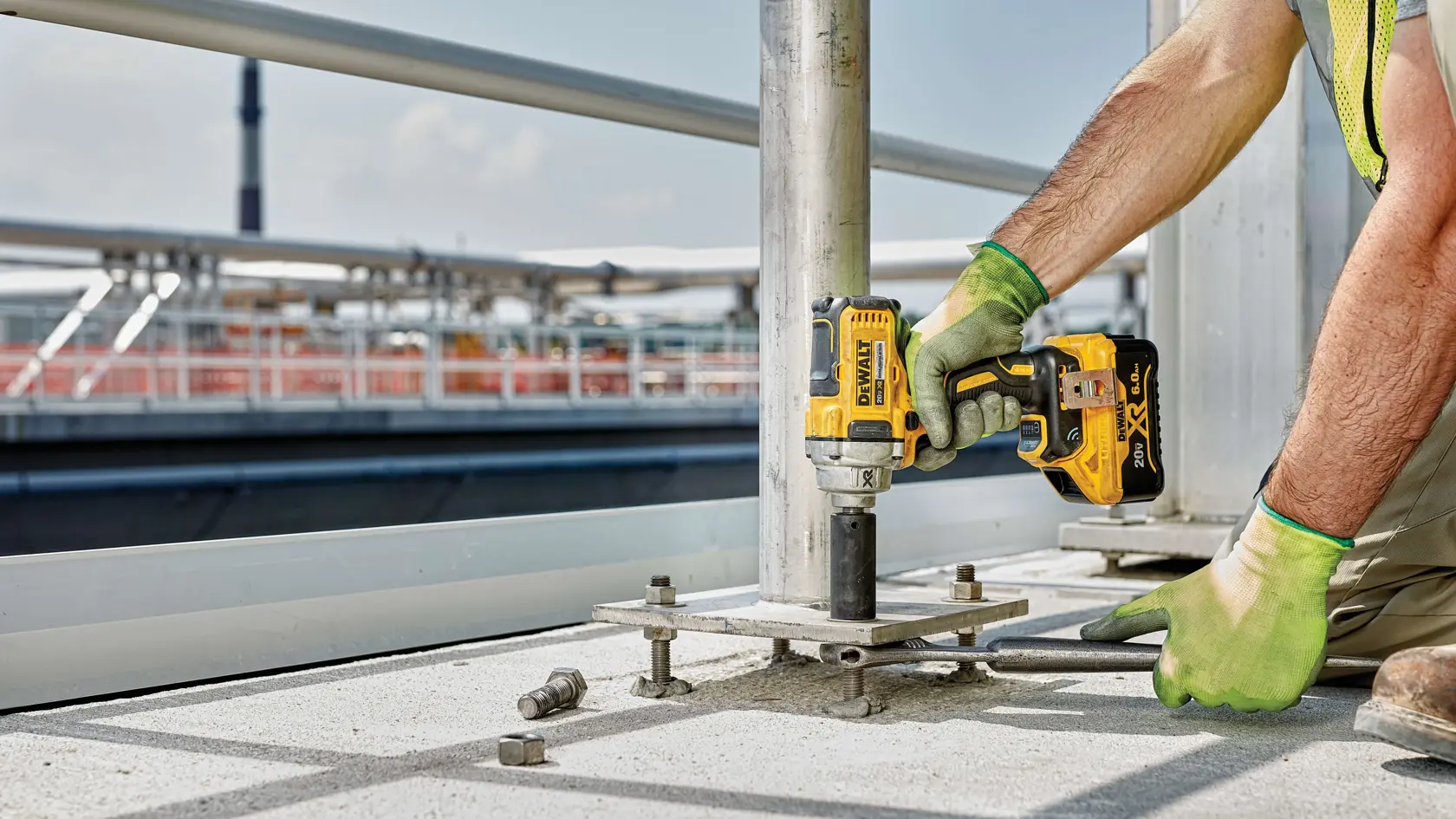 A person wearing green work gloves uses a yellow and black DEWALT cordless impact wrench to secure a metal pole to a concrete surface outdoors.