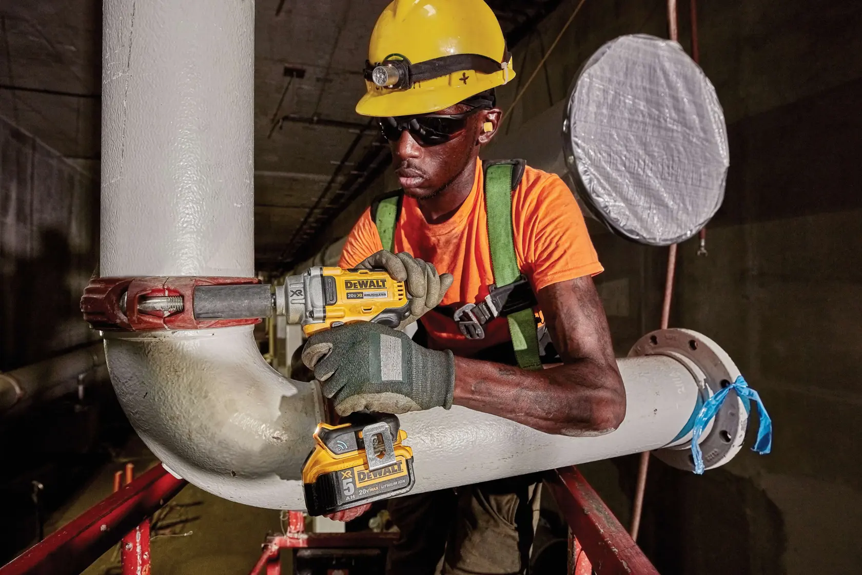 A worker wearing a yellow hard hat, gloves, and a safety harness is using a DEWALT DCF896P2 cordless impact wrench to work on a large industrial pipe in a dimly lit area.