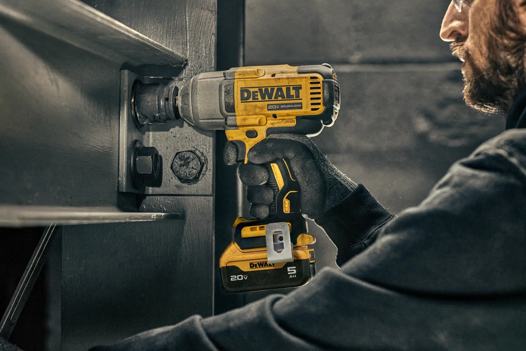 A person wearing gloves uses a DEWALT 20V cordless impact wrench to fasten a bolt on a large metal structure.