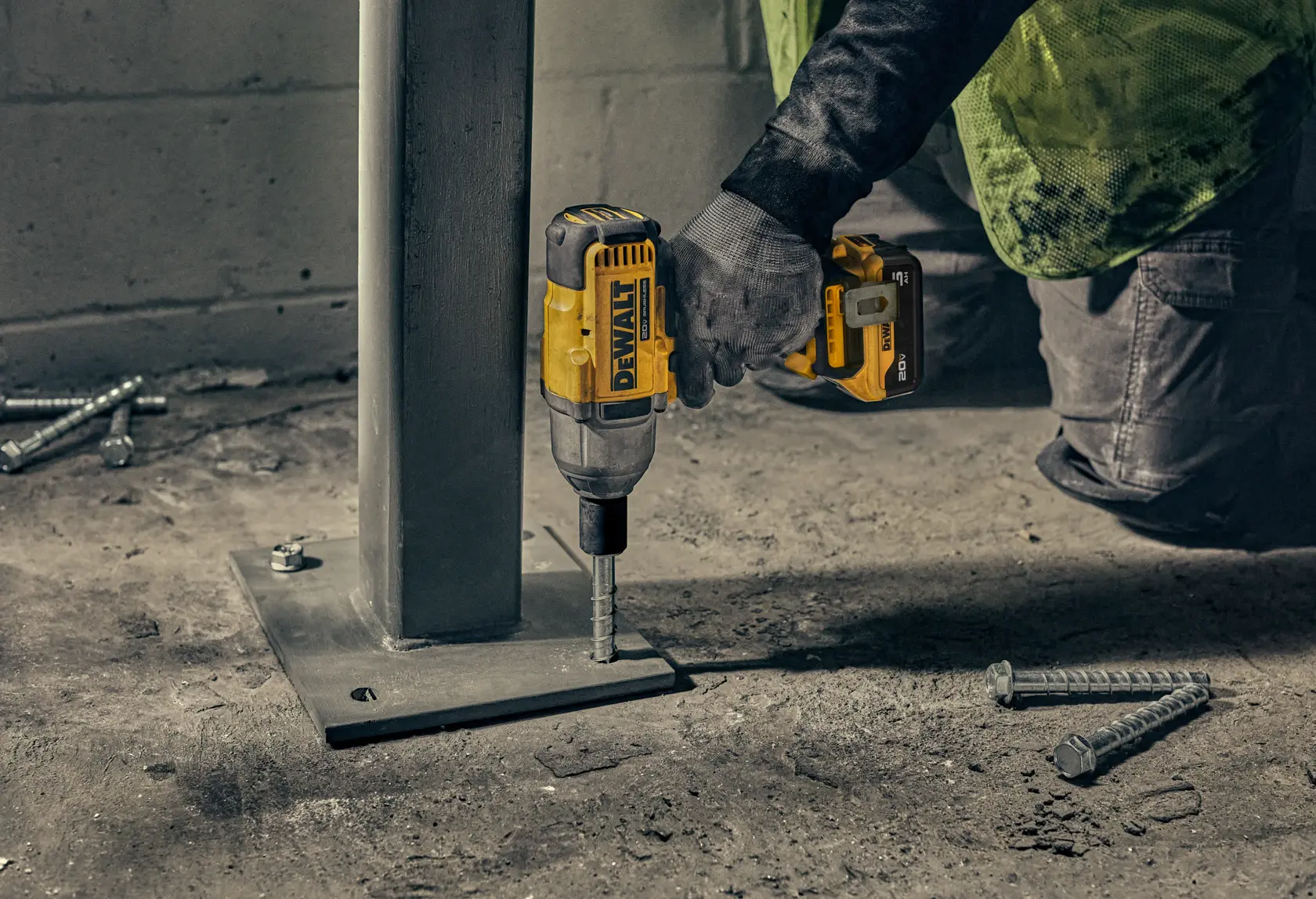 A person using a DEWALT cordless impact driver to secure a bolt at the base of a metal column. Several bolts are visible on the ground nearby, and the person is wearing gloves and work pants.