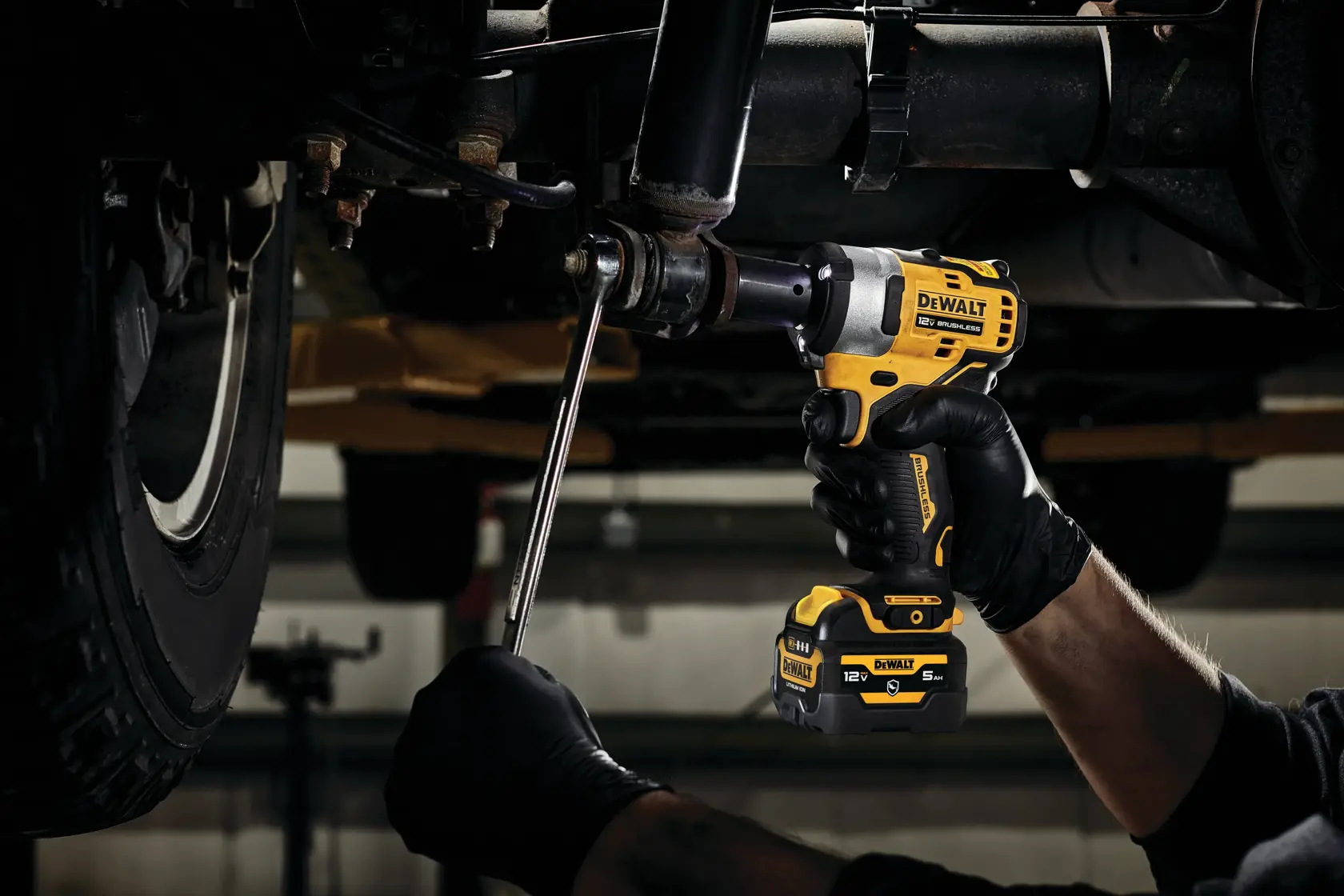 A person wearing black gloves is using a DEWALT DCF901B cordless impact wrench to work on a vehicle's undercarriage.