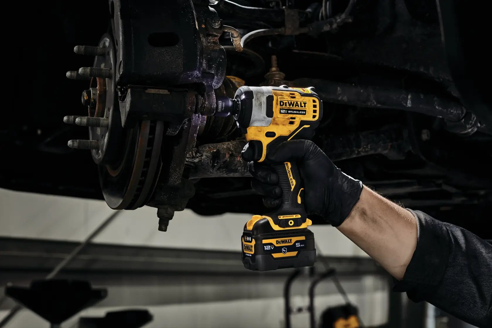 A person wearing a black glove uses a DEWALT 12V MAX cordless impact wrench to work on the undercarriage of a vehicle, focusing on a rusty brake assembly.