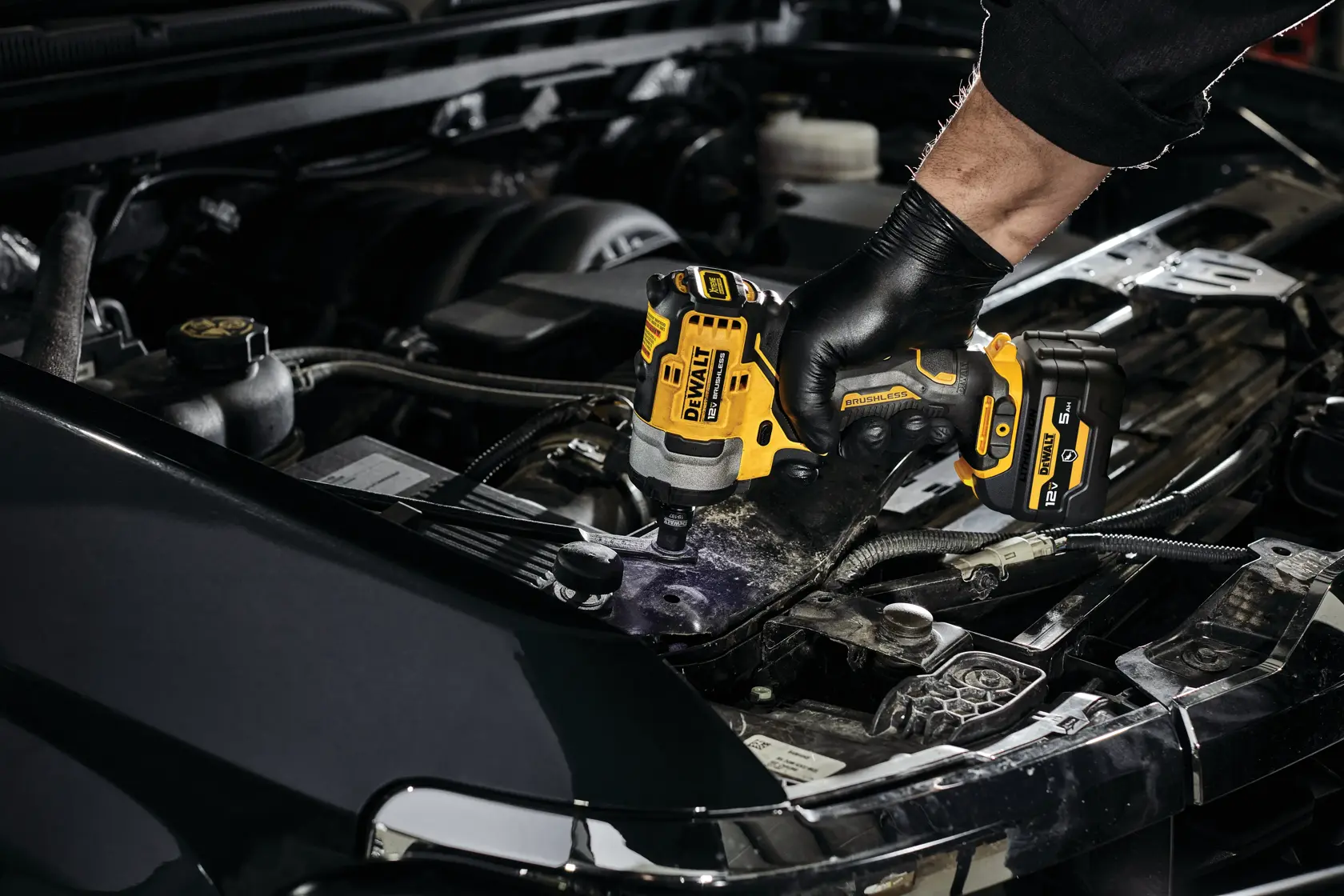 A person wearing black gloves uses a DEWALT cordless impact wrench, model DCF901GJ1G1, to work on a car engine in an automotive setting.