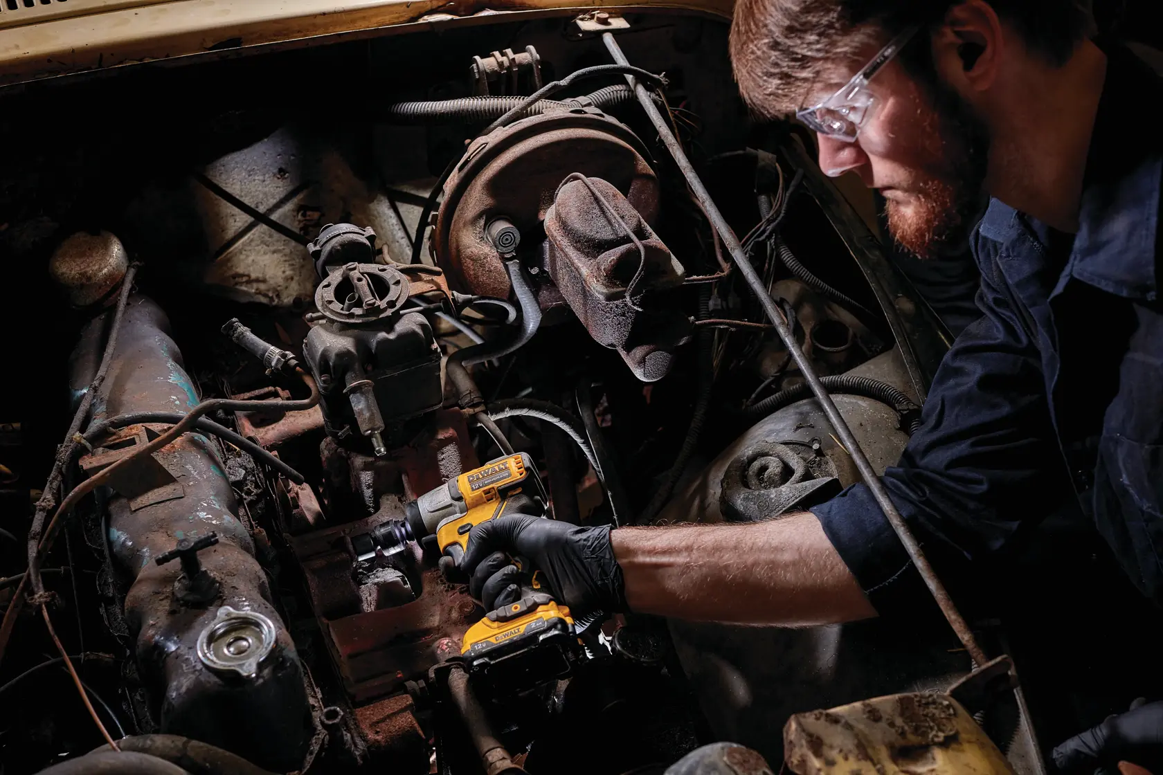 A person wearing gloves is using a DEWALT DCF902B cordless impact wrench to work on an engine inside a vehicle. The person's face is obscured and the focus is on the tool and the engine components.