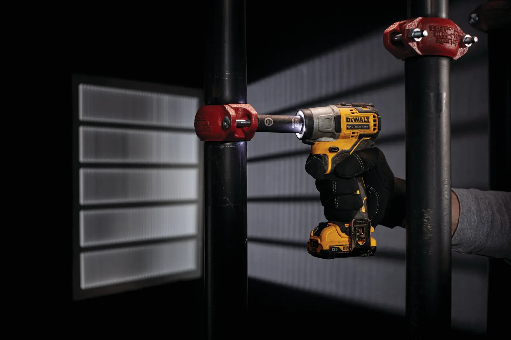 A person wearing a glove uses a DEWALT cordless impact driver to work on a pipe fitting. The tool is yellow and black, and the scene is indoors with dramatic lighting.