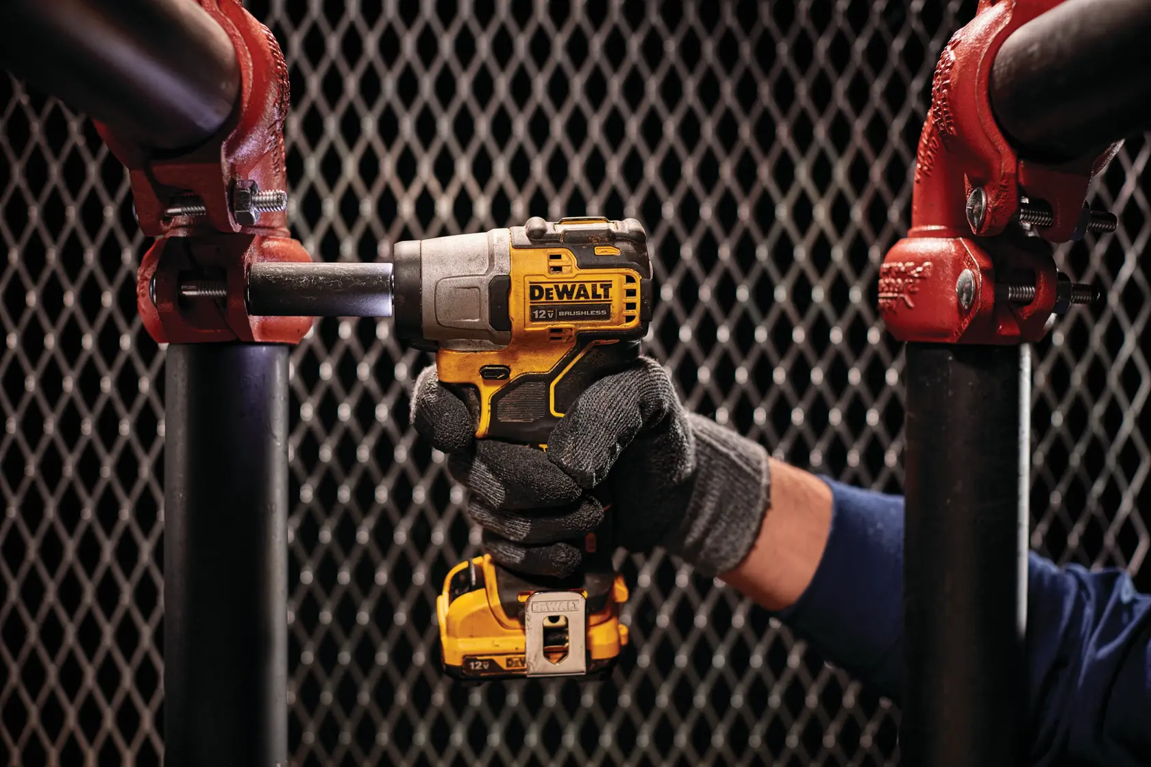 A person wearing work gloves uses a yellow and black DEWALT cordless impact wrench to tighten a bolt on a black metal pipe joint with red fittings. The background features a metal mesh pattern.