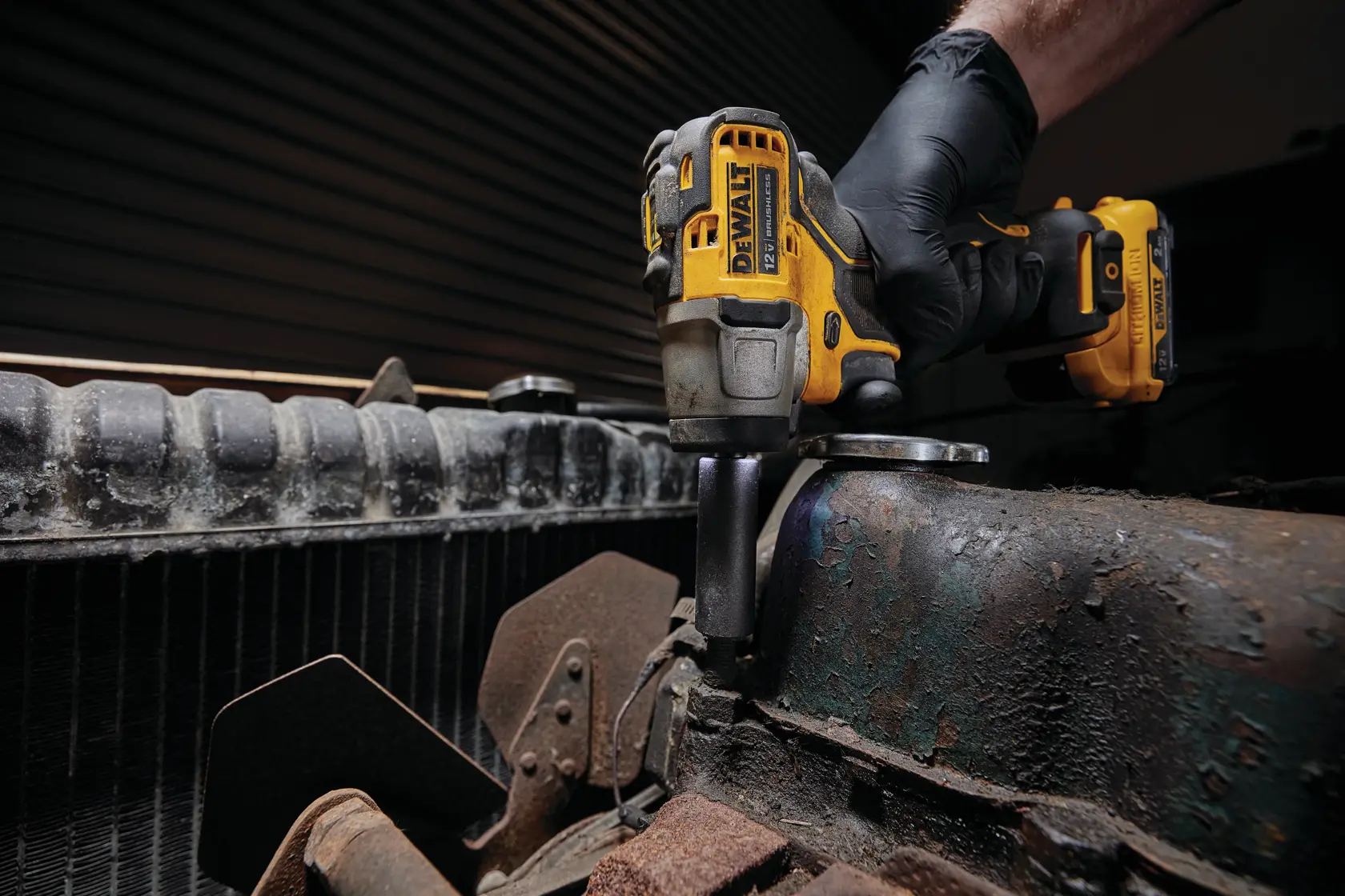 A person wearing a black glove is using a DEWALT cordless impact driver to work on a rusty engine or mechanical part.