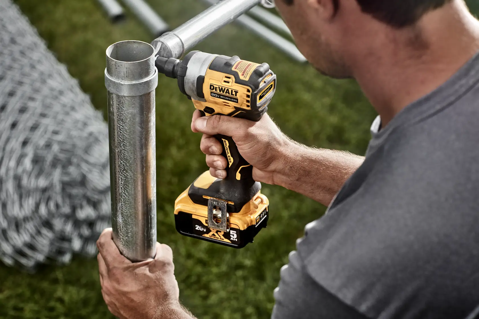 A person uses a DEWALT DCF911B cordless impact wrench to fasten a metal pipe outdoors. The tool is yellow and black, and the scene includes a coil of wire and green grass.