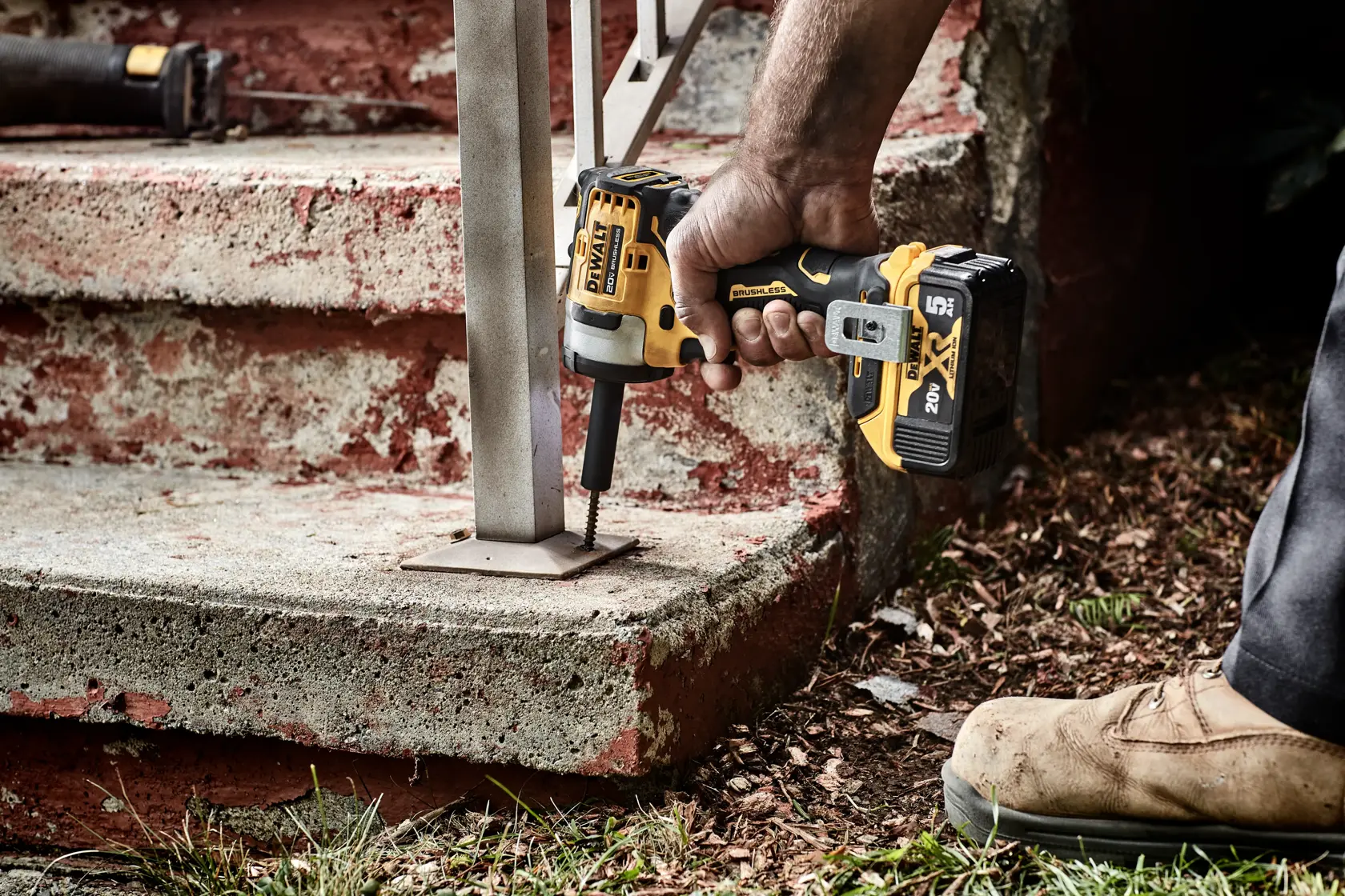 A person uses a DEWALT cordless impact driver to secure a metal handrail base to a concrete step outdoors.