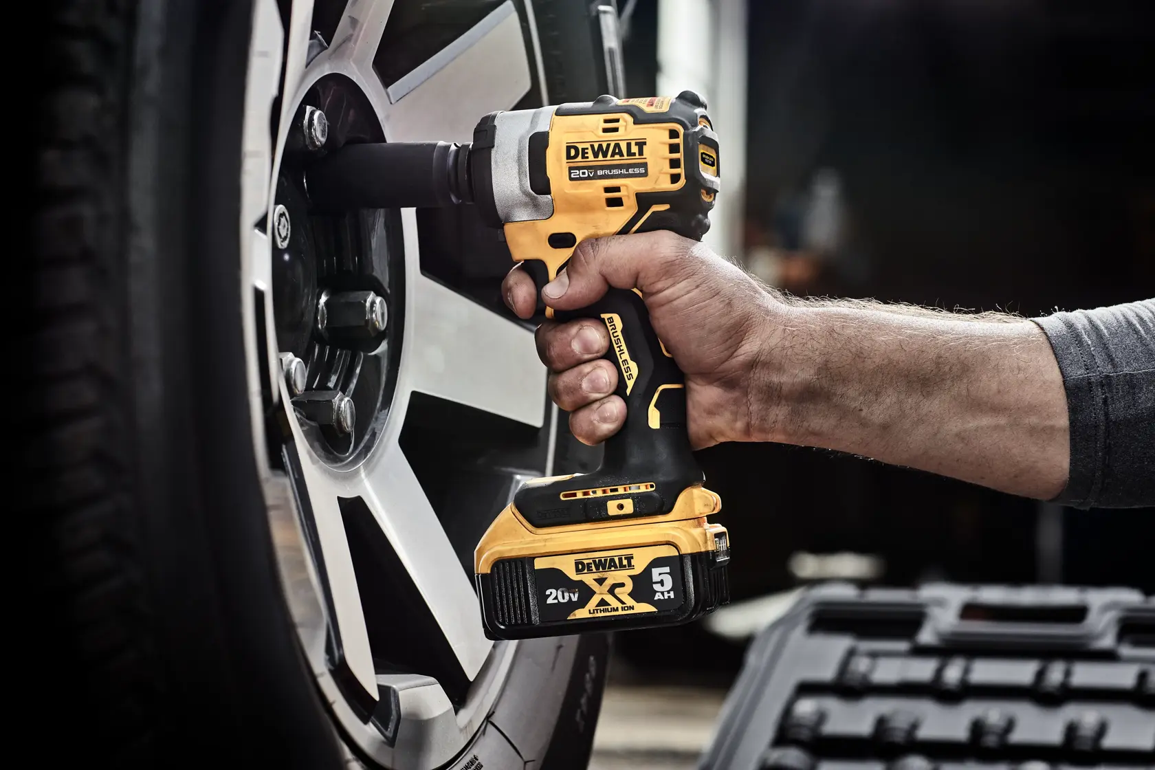 A hand using a DEWALT 20V cordless impact wrench to loosen or tighten a lug nut on a car wheel. The image highlights the oil-resistant feature of the tool's glass-filled nylon housing, which resists harsh automotive chemicals and solvents. The battery is not included.