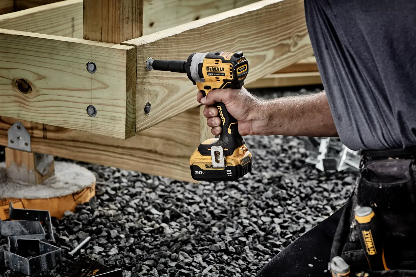 A person uses a DEWALT cordless impact wrench to fasten a bolt onto a wooden structure, likely during construction work on gravel ground.