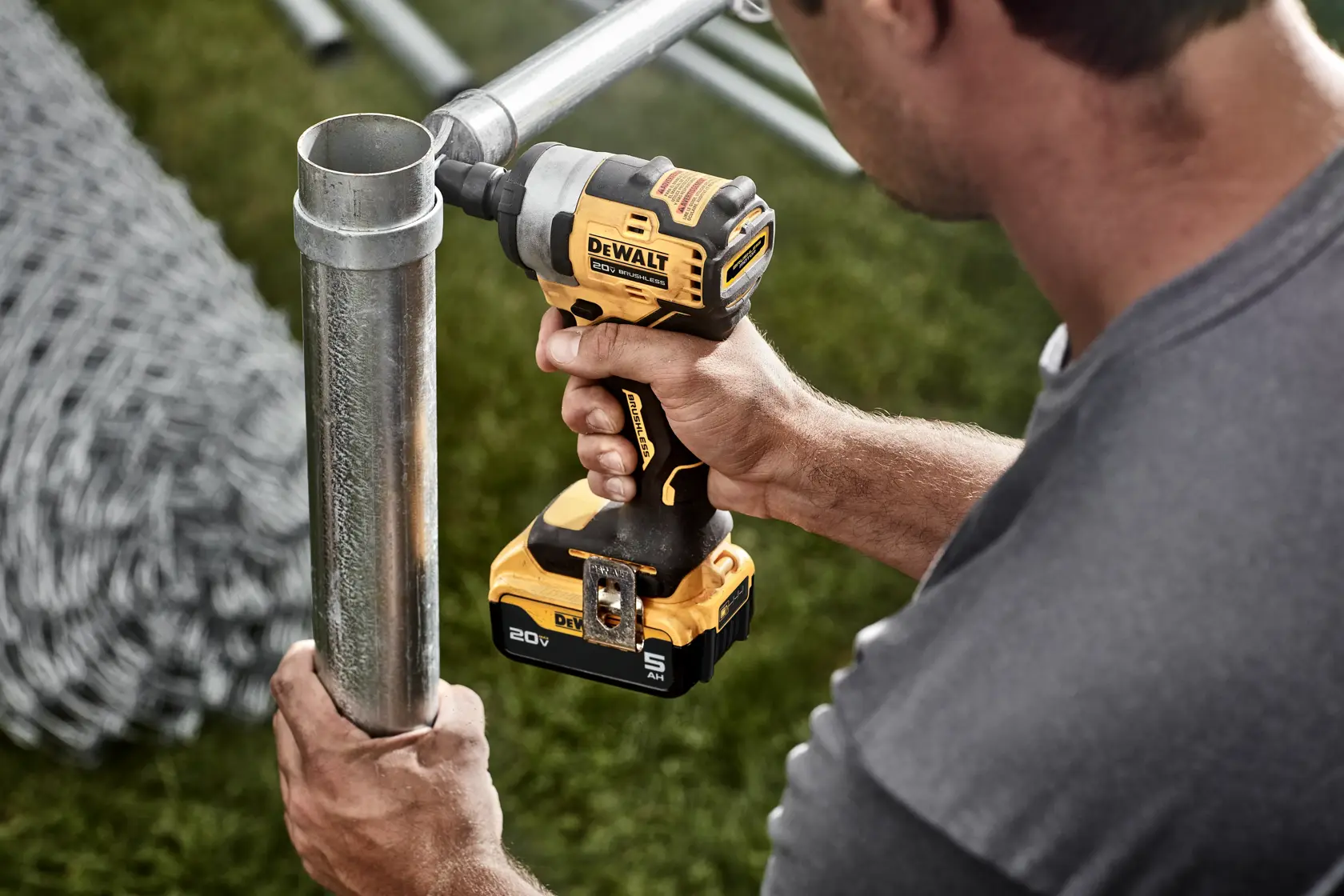 A person is using a DEWALT DCF911P2 cordless impact wrench to work on a metal pipe outdoors. The tool features a yellow and black color scheme and is powered by a 20V battery.