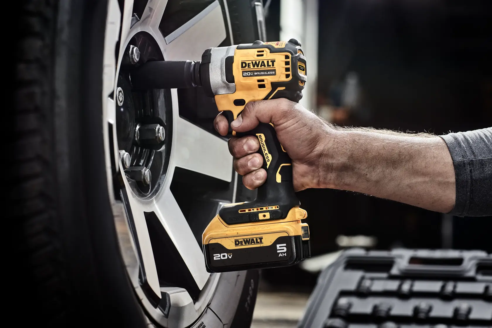 A DEWALT 20V brushless cordless impact wrench being used to loosen a lug nut on a car wheel. The product's oil-resistant feature and glass-filled nylon housing, which resists effects of harsh automotive chemicals and solvents, are highlighted in the text.