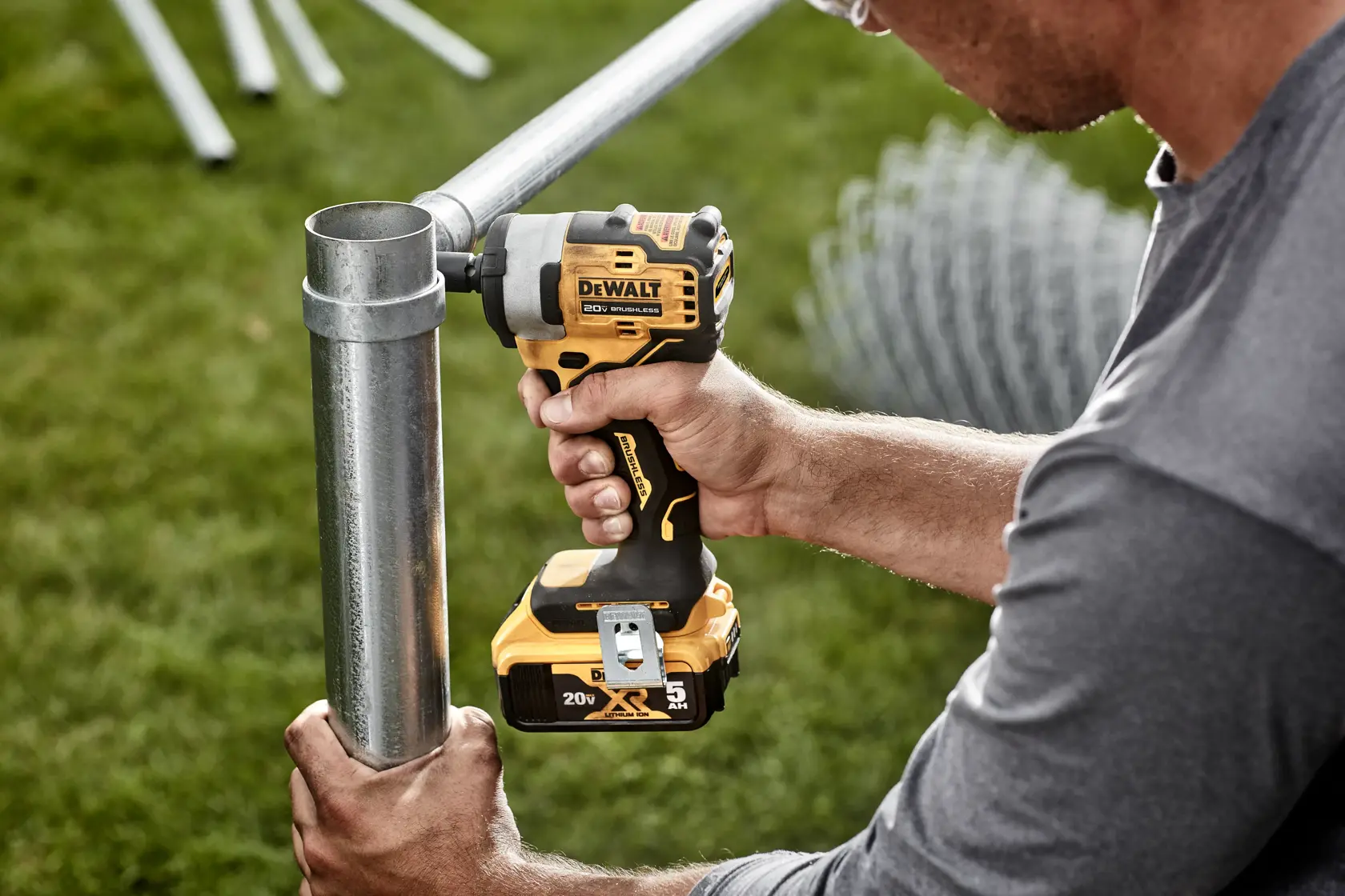 A person using a DEWALT 20V cordless impact wrench (model DCF913B) to tighten a metal pipe outdoors, with green grass in the background.