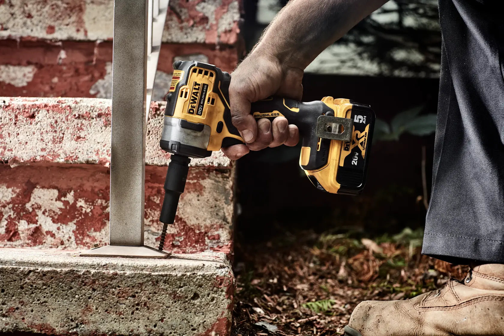 A person is using a DEWALT cordless impact wrench to drive a screw into a metal base attached to a concrete step. The tool is yellow and black and held in the right hand. The person is wearing work boots and dark pants.