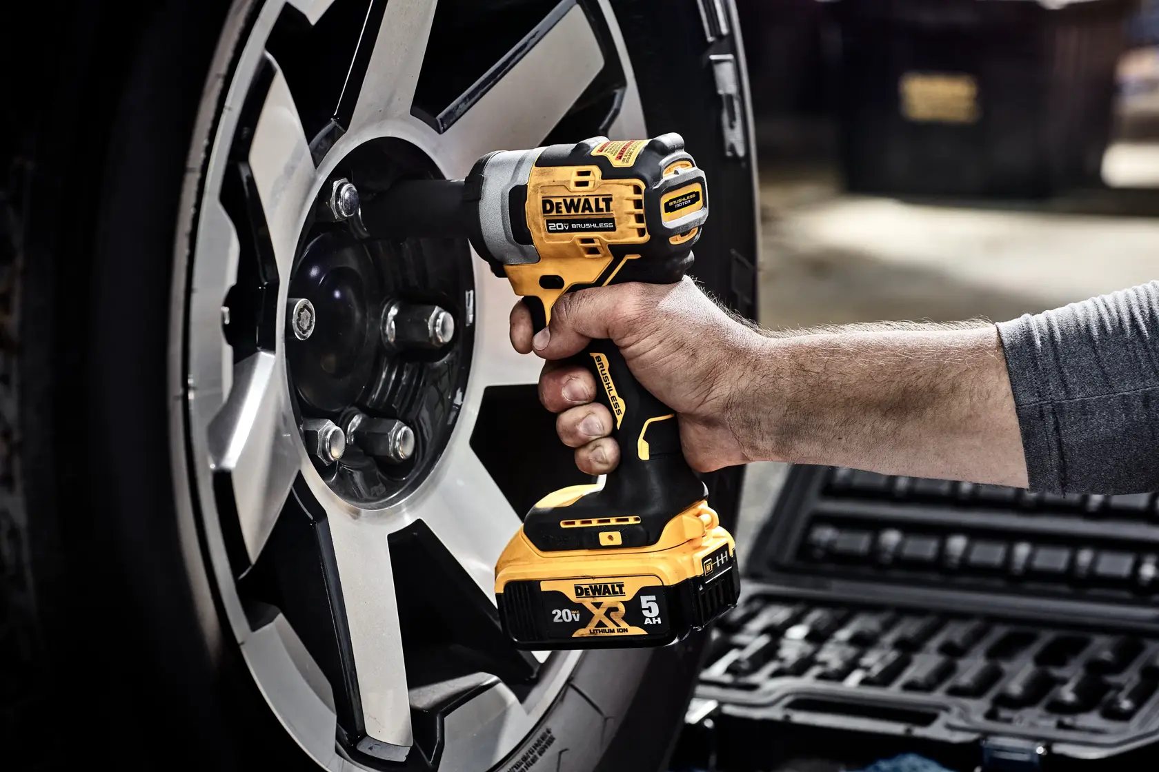 A person is using a DEWALT 20V cordless impact wrench to remove a lug nut from a car wheel. The tool is highlighted for its oil-resistant, glass-filled nylon housing, which resists harsh automotive chemicals and solvents. Text notes that the battery is not included.