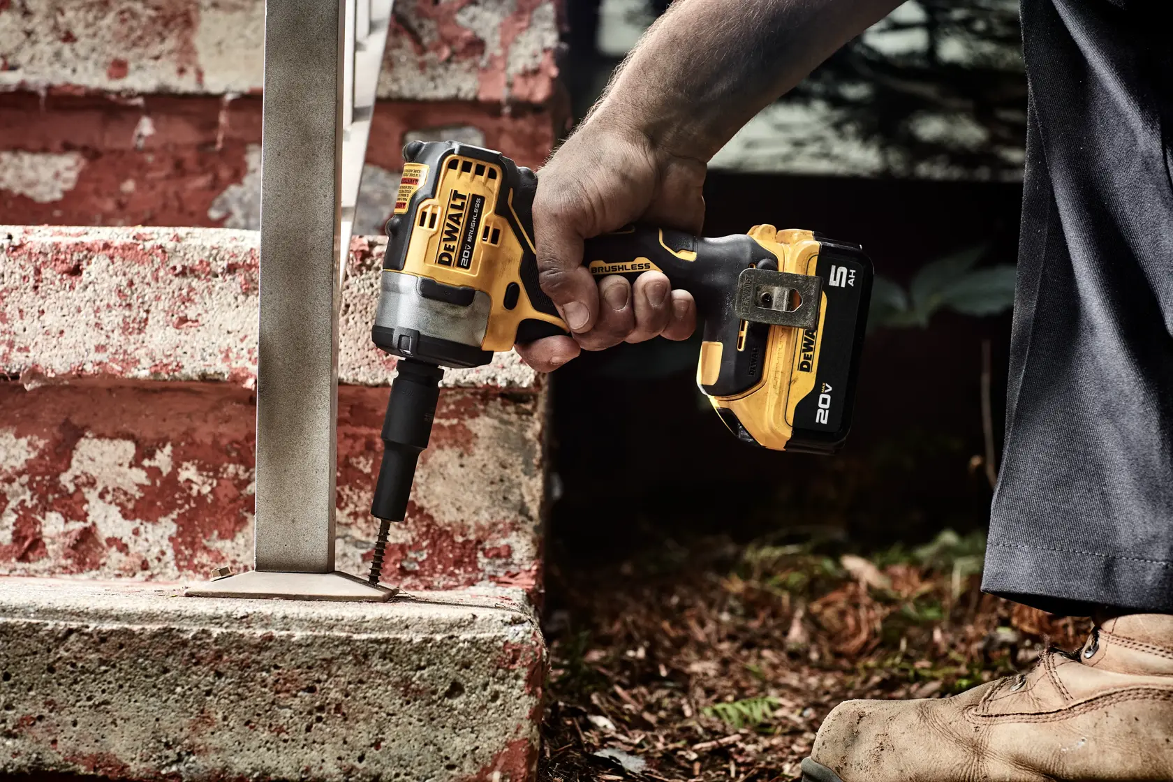 A person uses a DEWALT cordless impact wrench to drive a screw into a metal bracket attached to a concrete step. The tool is yellow and black, and the person is wearing work boots.