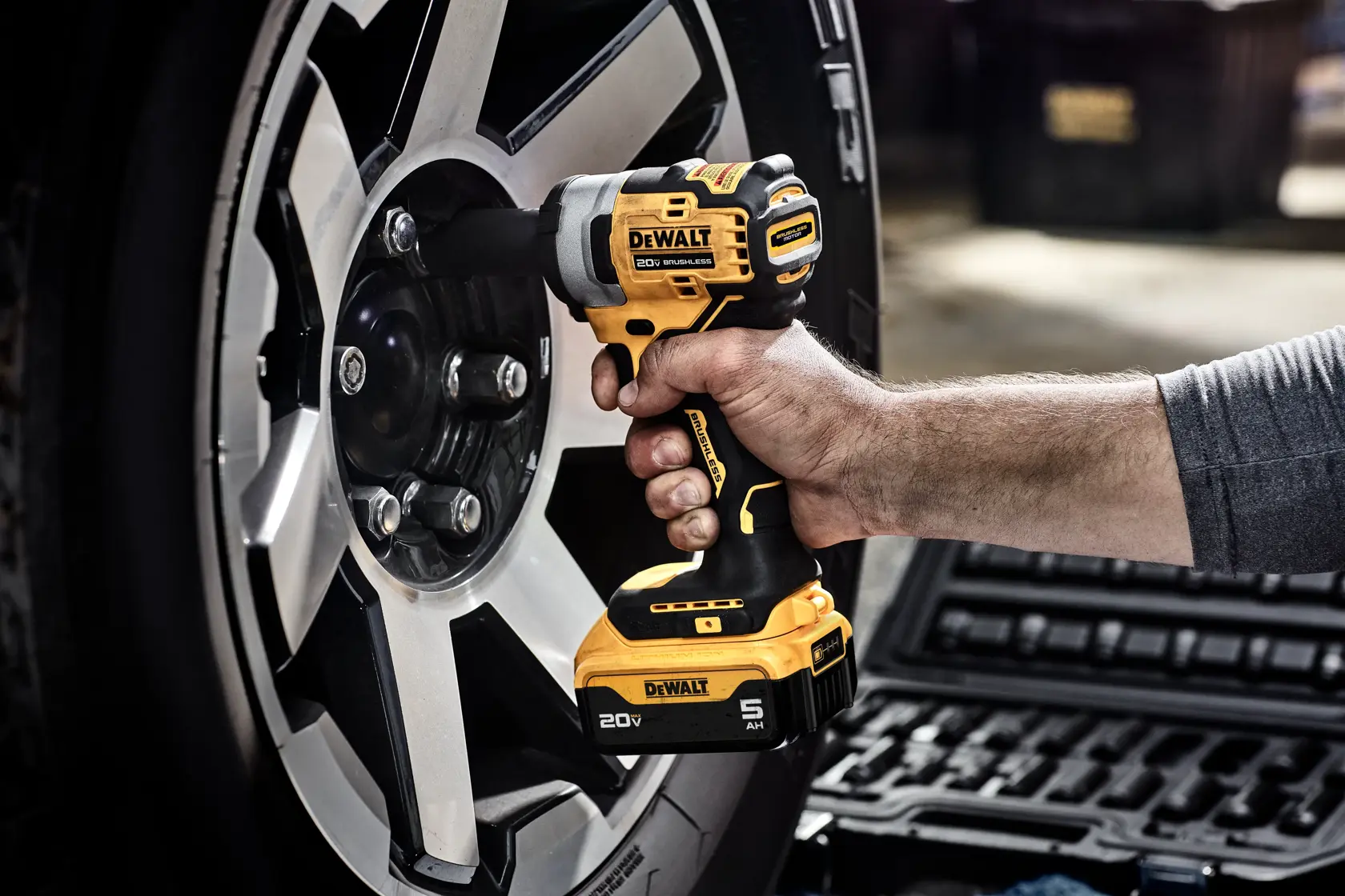 A person using a DEWALT 20V cordless impact wrench to tighten or loosen lug nuts on a car wheel. The product is described as oil-resistant, with glass-filled nylon housing that resists effects of harsh automotive chemicals and solvents.
