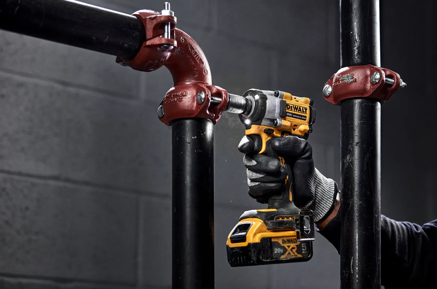 A person wearing a work glove uses a DEWALT cordless impact wrench to tighten a metal pipe fitting. The tool is yellow and black and is being used in an industrial setting with black pipes and red connectors.