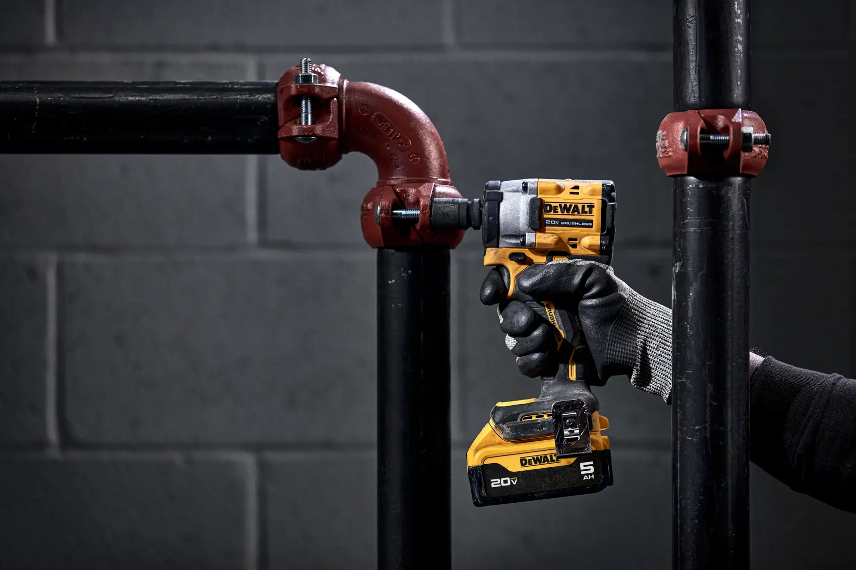 A person wearing protective gloves uses a yellow and black DEWALT cordless impact wrench (model DCF921P2) to work on metal pipe fittings in an industrial setting.