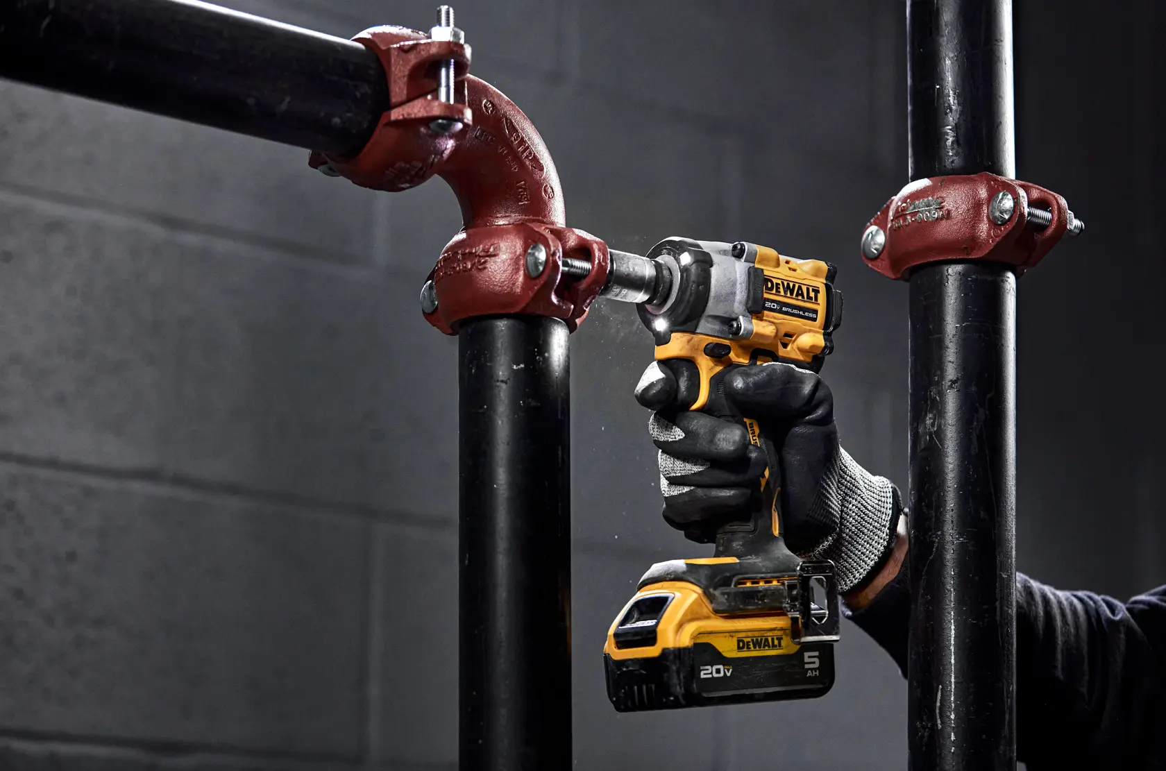 A gloved hand uses a yellow and black DEWALT 20V cordless impact wrench (model DCF921P2) to tighten a metal pipe fitting in an industrial setting.