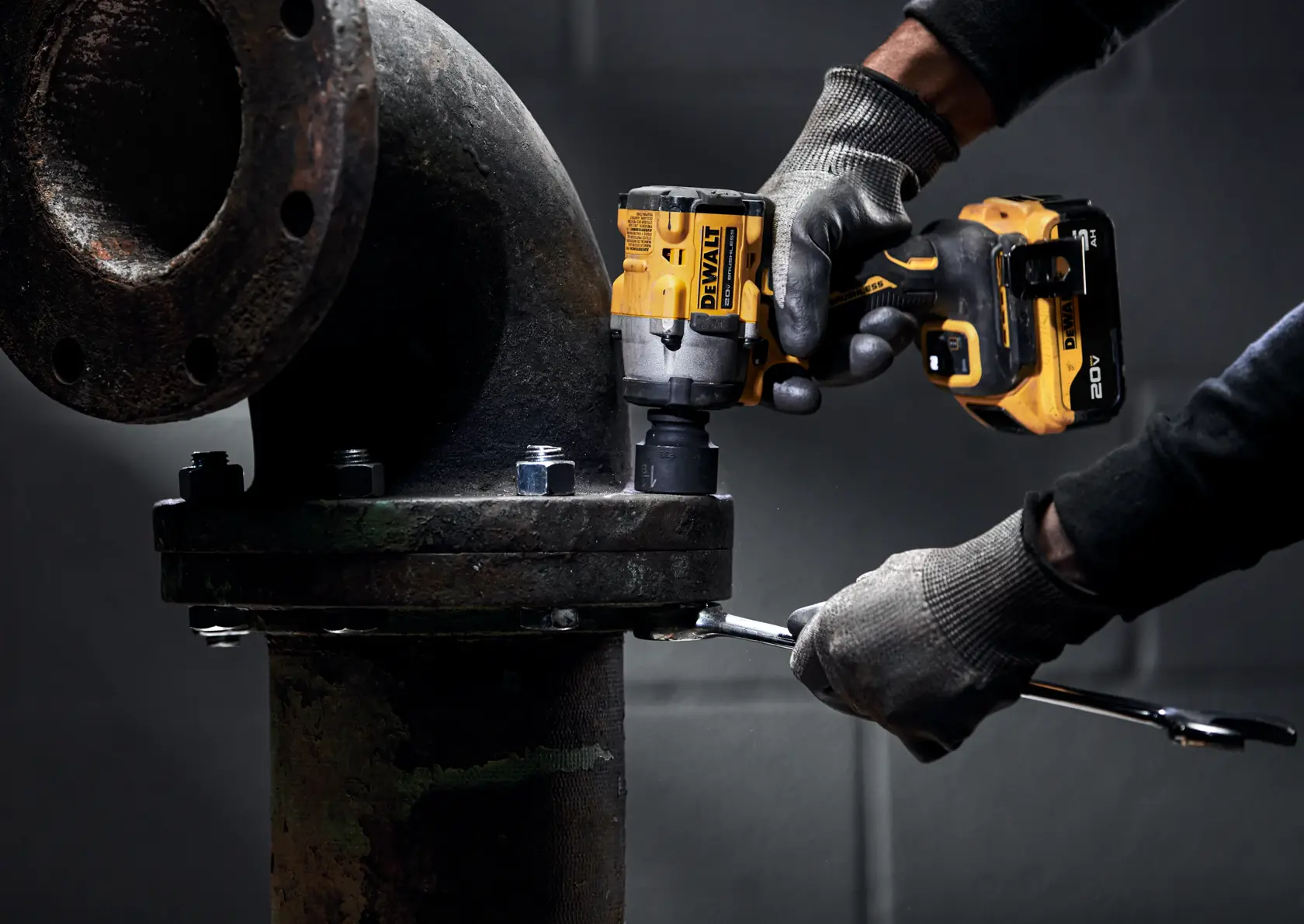 A person wearing gloves is using a DEWALT cordless impact wrench to tighten bolts on a large industrial pipe fitting. The yellow and black power tool is held firmly, while another hand uses a wrench on the bolt.