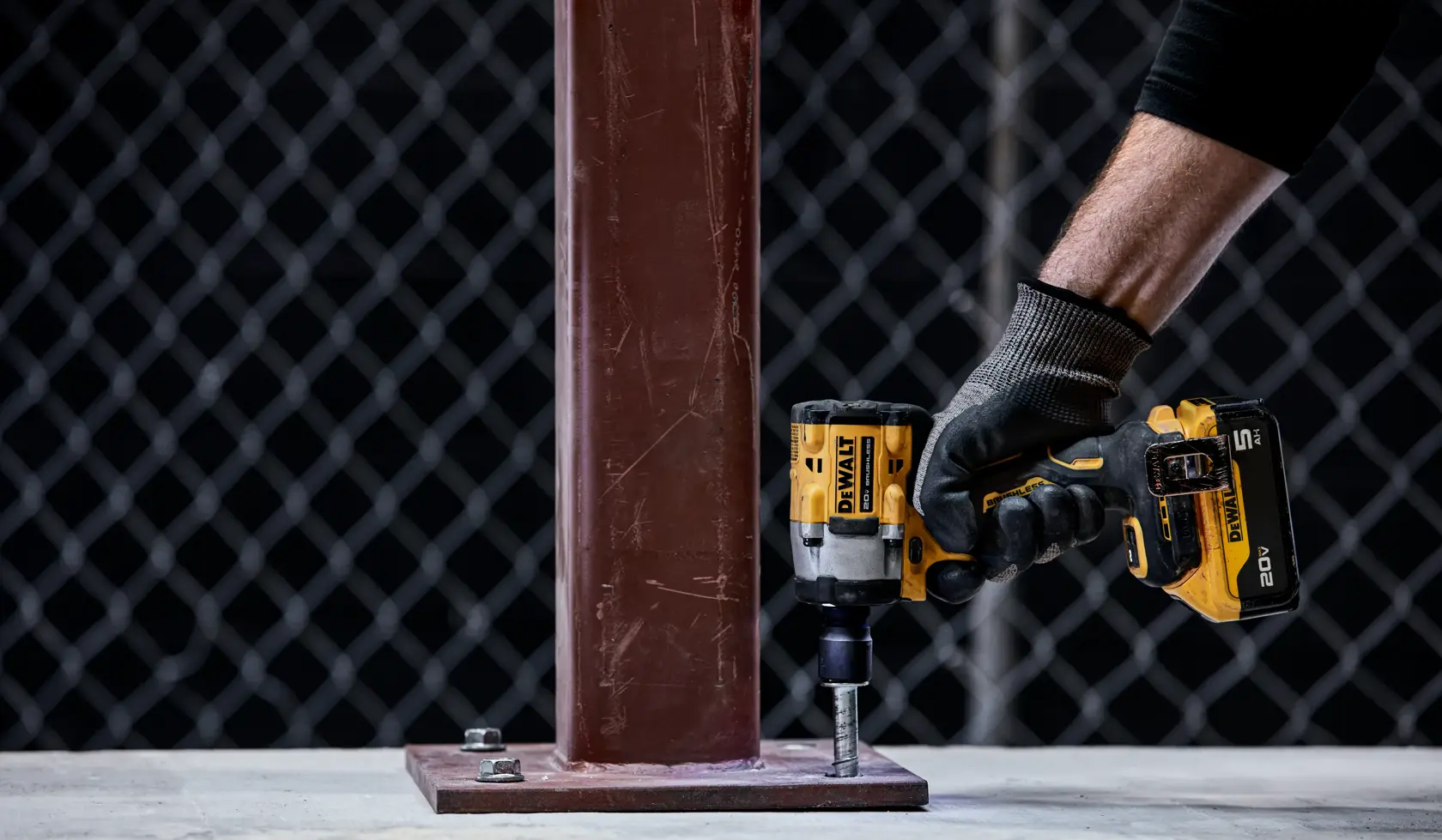 A person wearing a black sleeve and work gloves uses a DEWALT cordless impact wrench (model DCF921P2) to tighten a bolt on a metal base plate attached to a steel beam. The scene is set in an industrial area with a chain-link fence in the background.