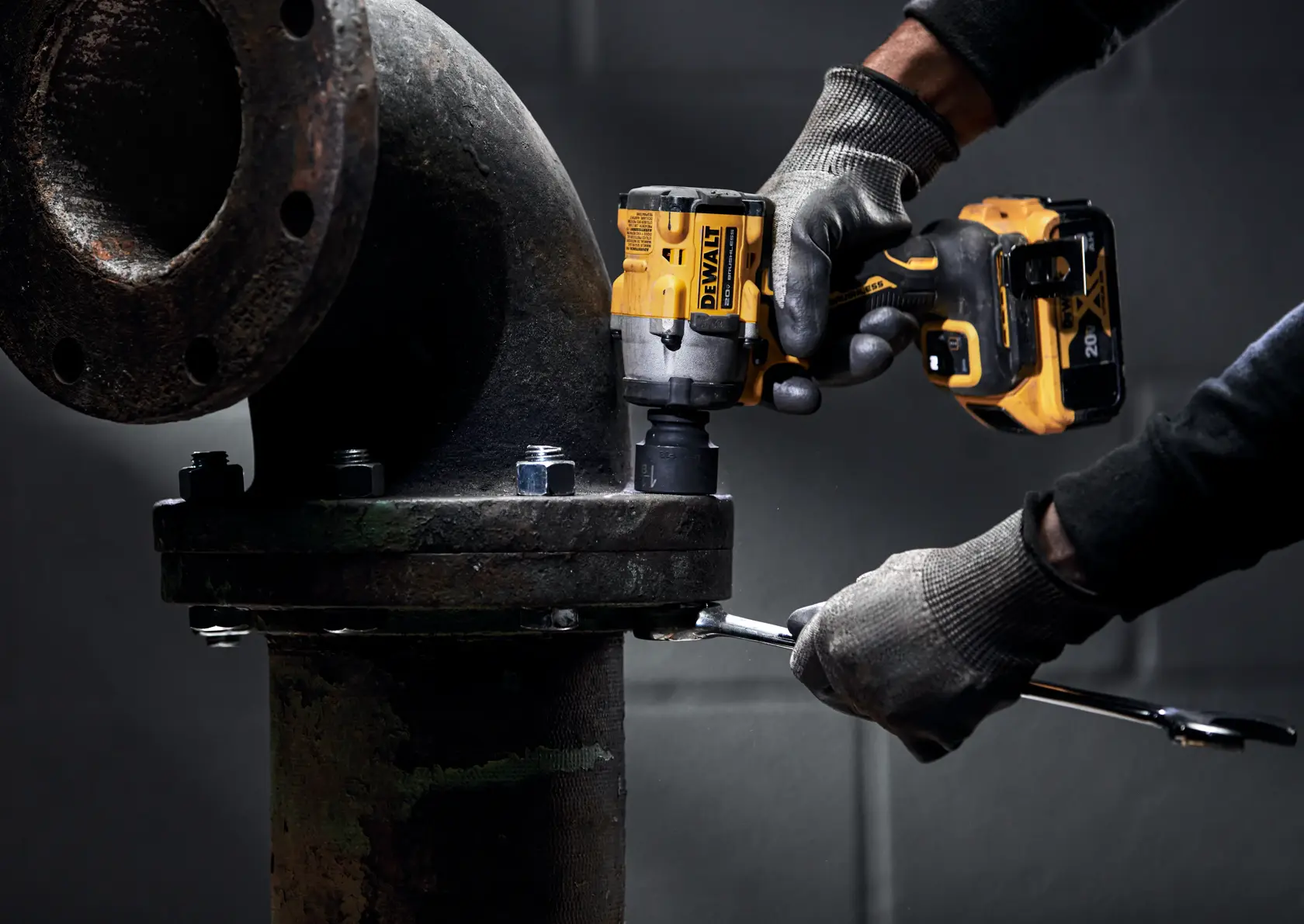 A person wearing gloves uses a DEWALT cordless impact wrench to tighten bolts on a large industrial pipe flange. The scene is set in a workshop or industrial setting.