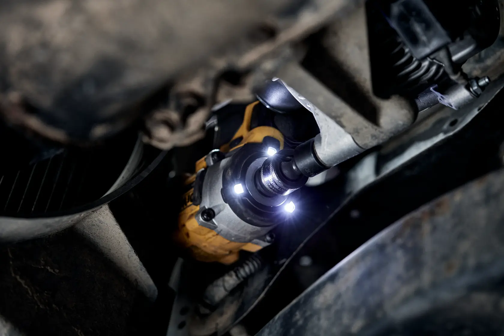 A close-up view of a DEWALT cordless impact wrench being used on a vehicle part in a dark, confined space, with its LED lights illuminating the work area.