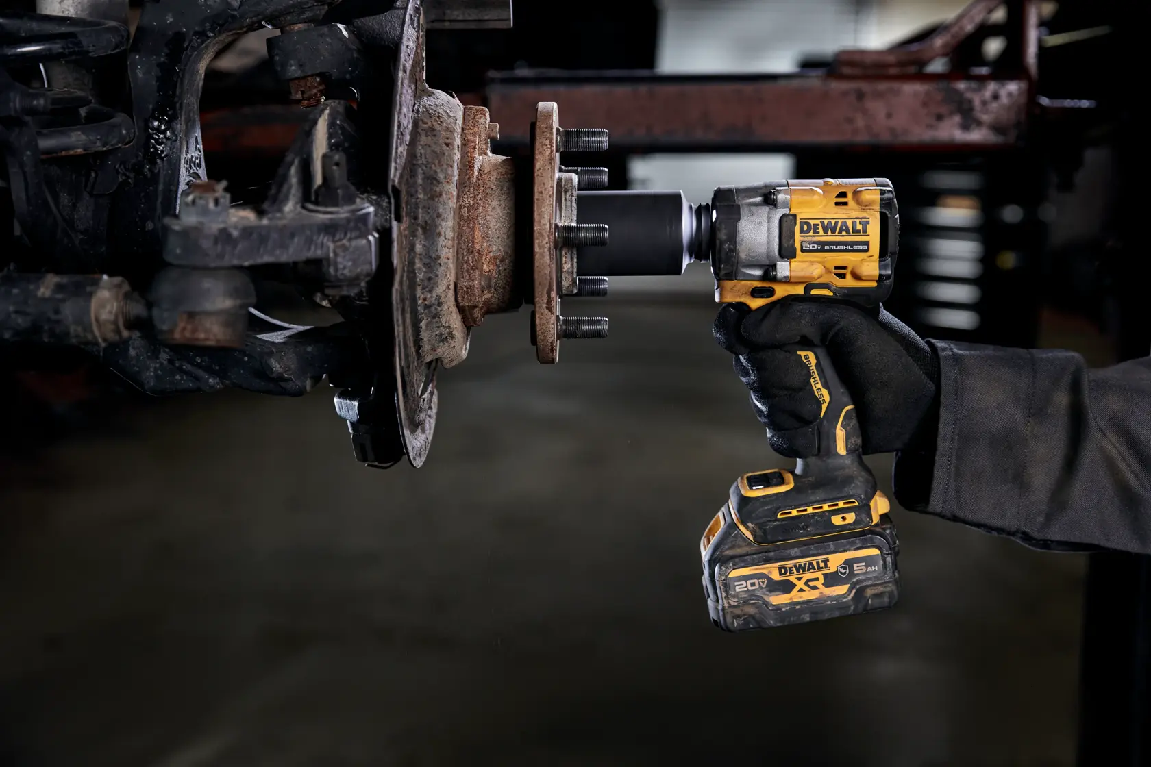 A person wearing a glove uses a DEWALT cordless impact wrench to work on a rusty vehicle part, likely a wheel hub or axle assembly, in a garage or workshop setting.