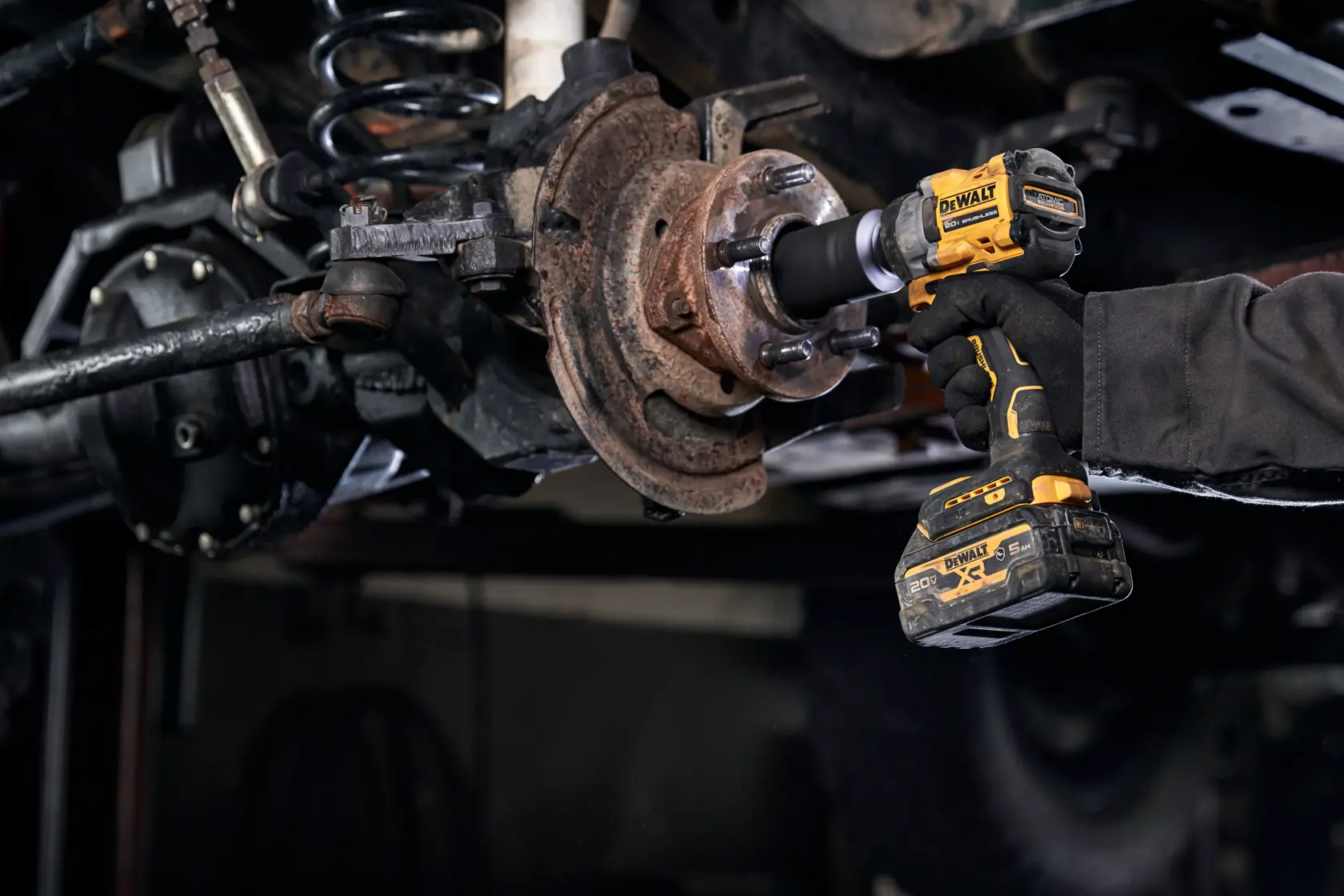 A person using a DEWALT DCF923B cordless impact wrench to work on an automotive part, possibly a wheel hub or brake assembly, in a workshop setting.