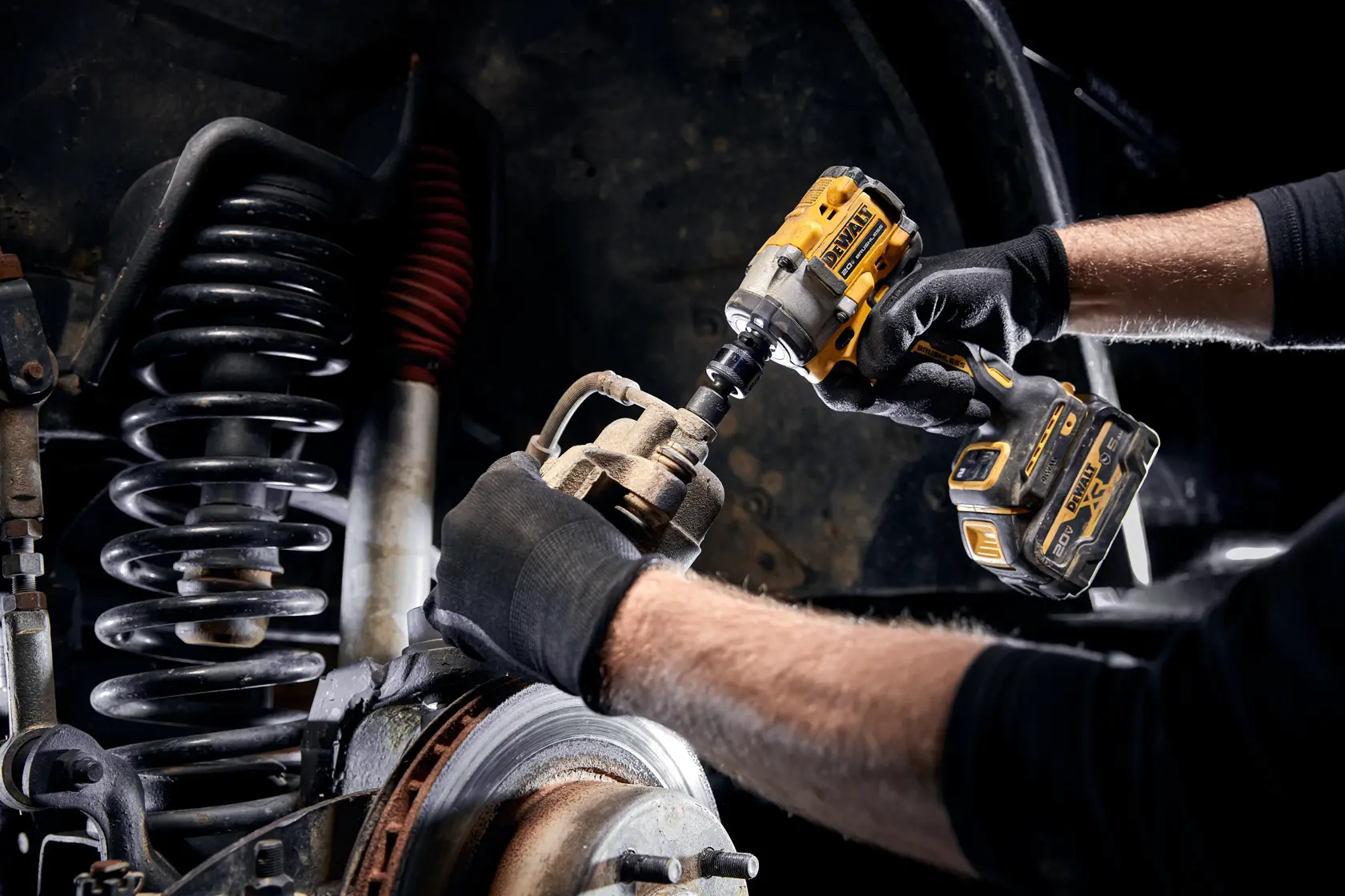 A person wearing black gloves uses a DEWALT cordless impact wrench to work on the brake assembly of a vehicle, with suspension components visible in the background.