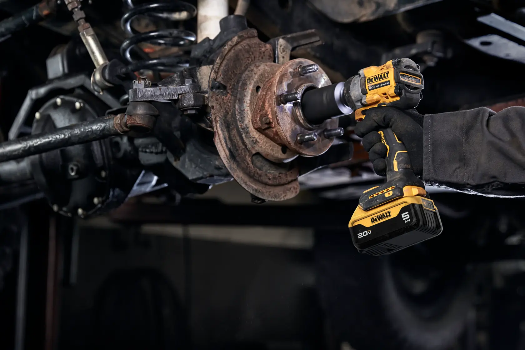A person using a DEWALT 20V cordless impact wrench to work on a rusty vehicle suspension component in a workshop setting.