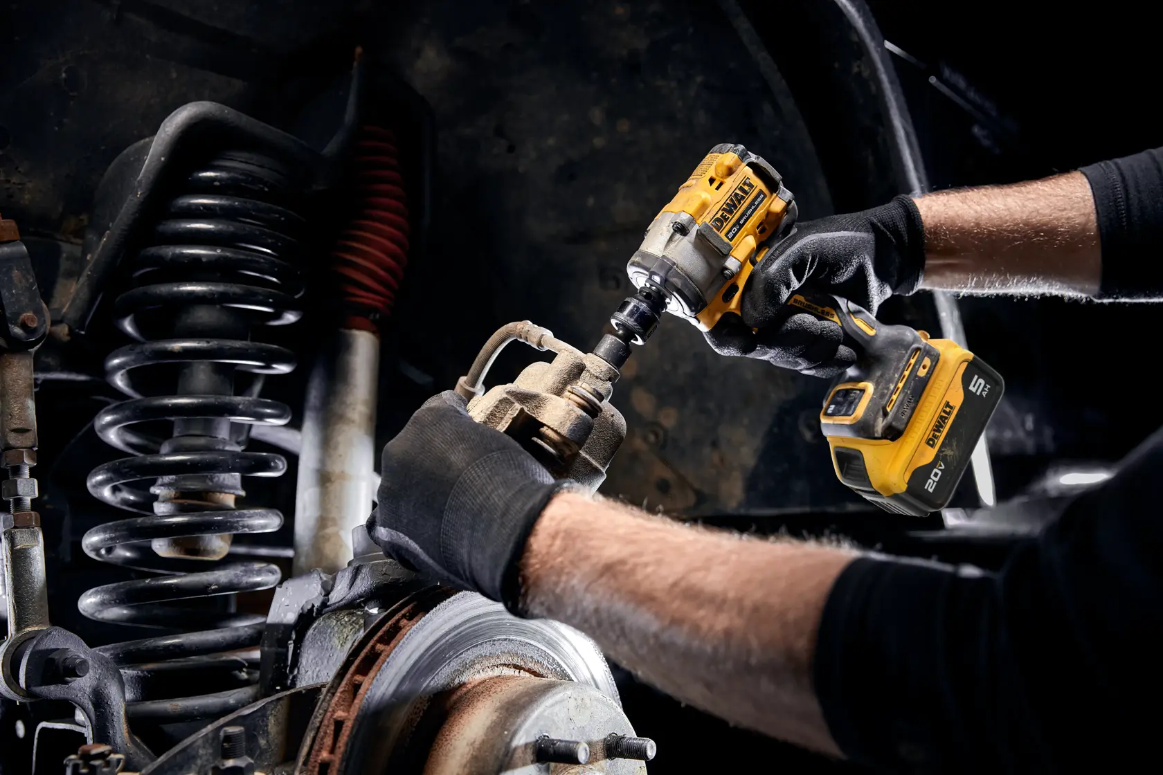 A person wearing black gloves is using a yellow DEWALT cordless impact wrench to work on a vehicle suspension and brake assembly.