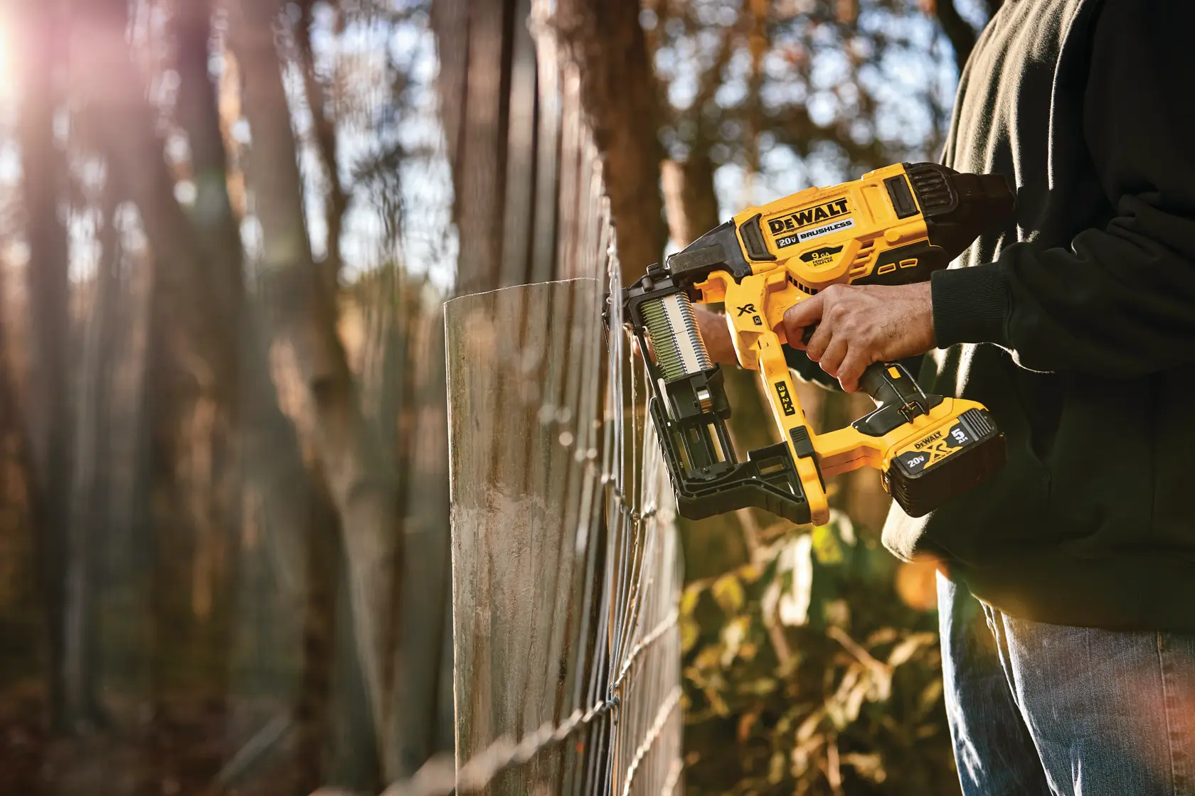 A person using a DEWALT cordless fencing stapler to secure wire fencing outdoors, with trees and sunlight in the background.
