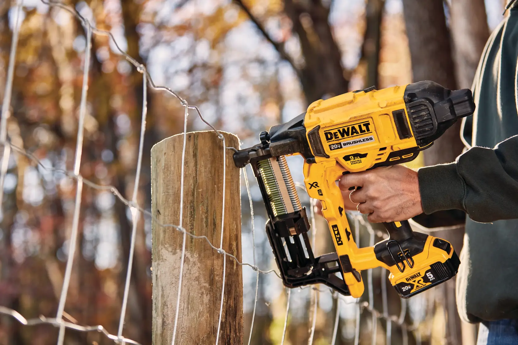A person using a DEWALT 20V cordless fencing stapler to secure wire fencing to a wooden post outdoors.