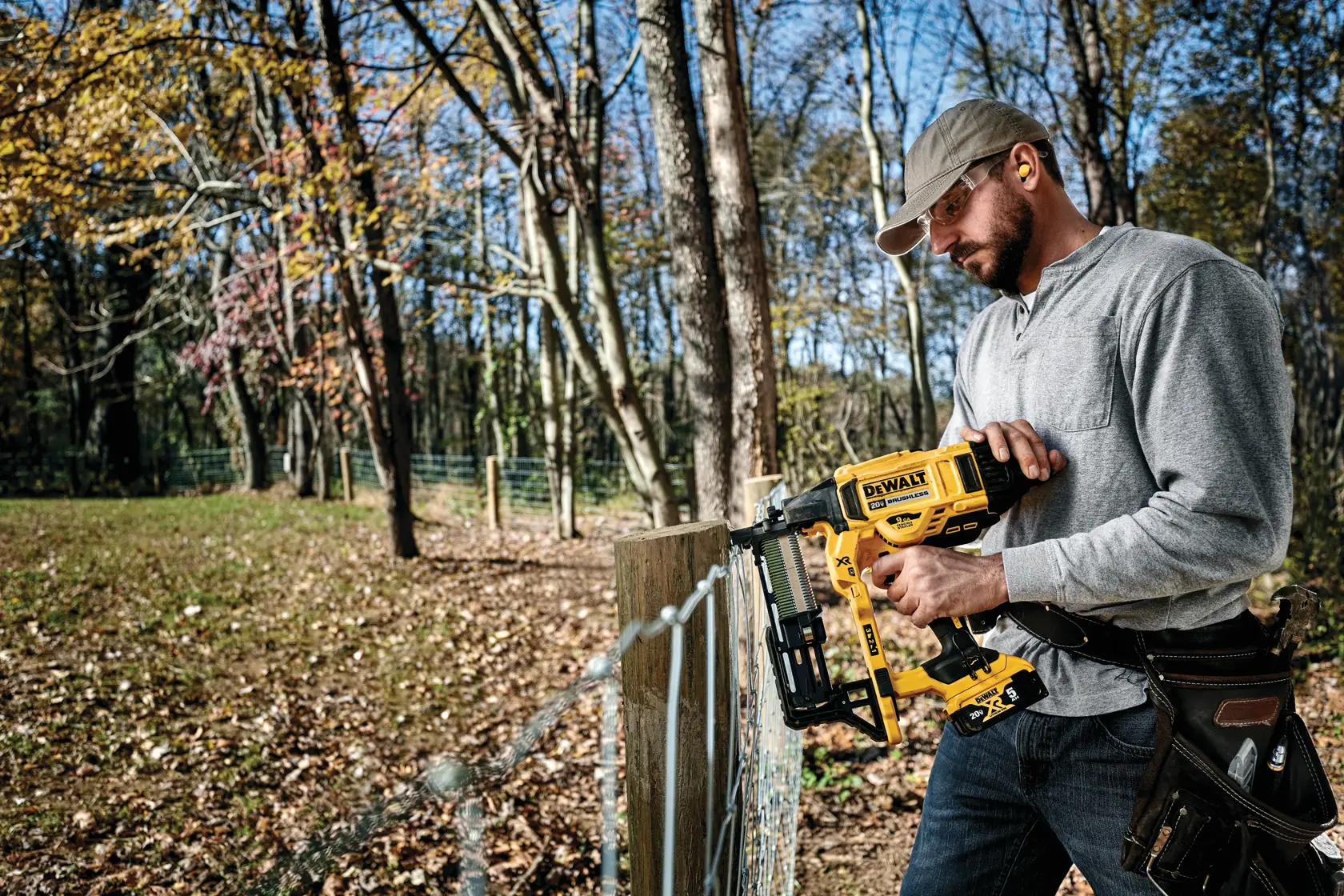 A person outdoors using a DEWALT DCFS950B cordless fencing stapler to secure wire mesh to a wooden fence post. The individual is wearing a gray long-sleeve shirt, a tool belt, and a cap, and their face is blurred. Trees and autumn foliage can be seen in the background.
