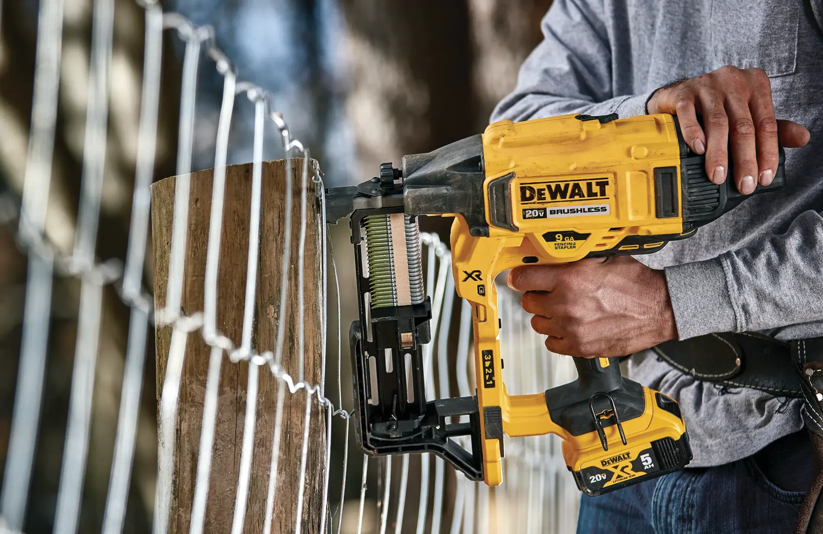 A person using a DEWALT 20V brushless cordless nailer to secure wire fencing to a wooden post outdoors.