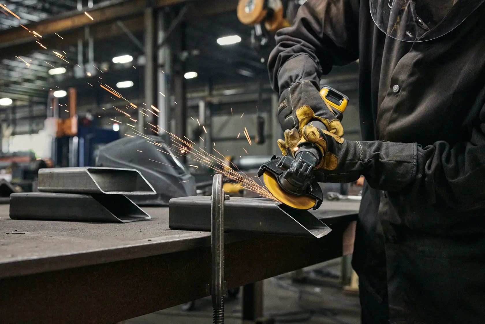 A person wearing safety gloves and protective clothing is using a DEWALT cordless angle grinder to grind a piece of metal on a workbench, producing sparks in an industrial workshop setting.