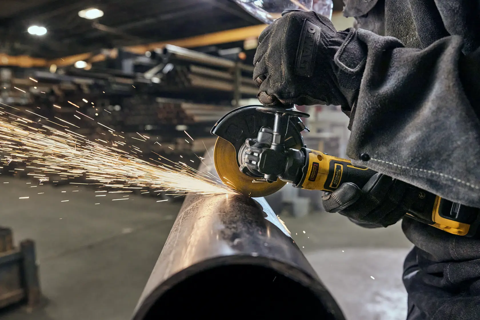 A person wearing protective gloves and clothing is using a DEWALT cordless angle grinder to cut through a metal pipe, creating sparks in a workshop setting.