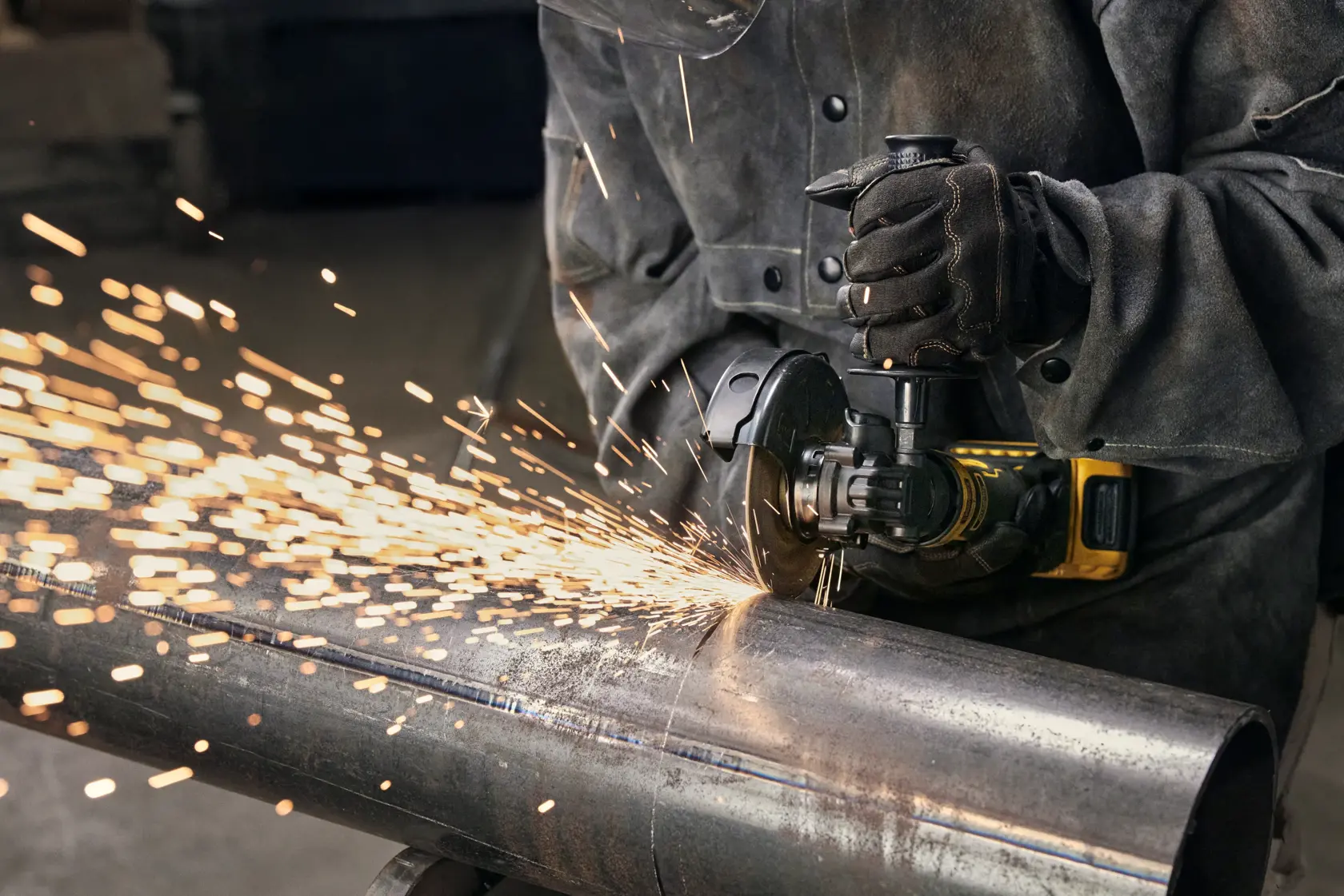 A person wearing protective clothing and gloves is using a DEWALT angle grinder to cut a metal pipe, with sparks flying as the tool operates.