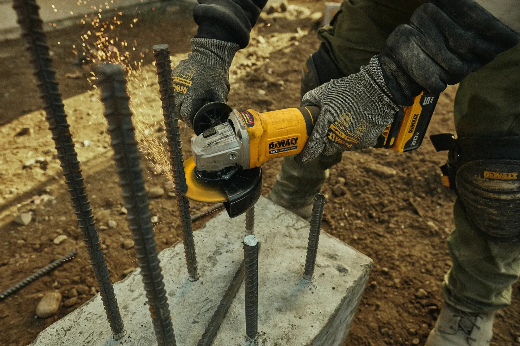 A person wearing gloves uses a yellow DEWALT cordless angle grinder to cut steel rebar protruding from a concrete block, with sparks visible during the cutting process.