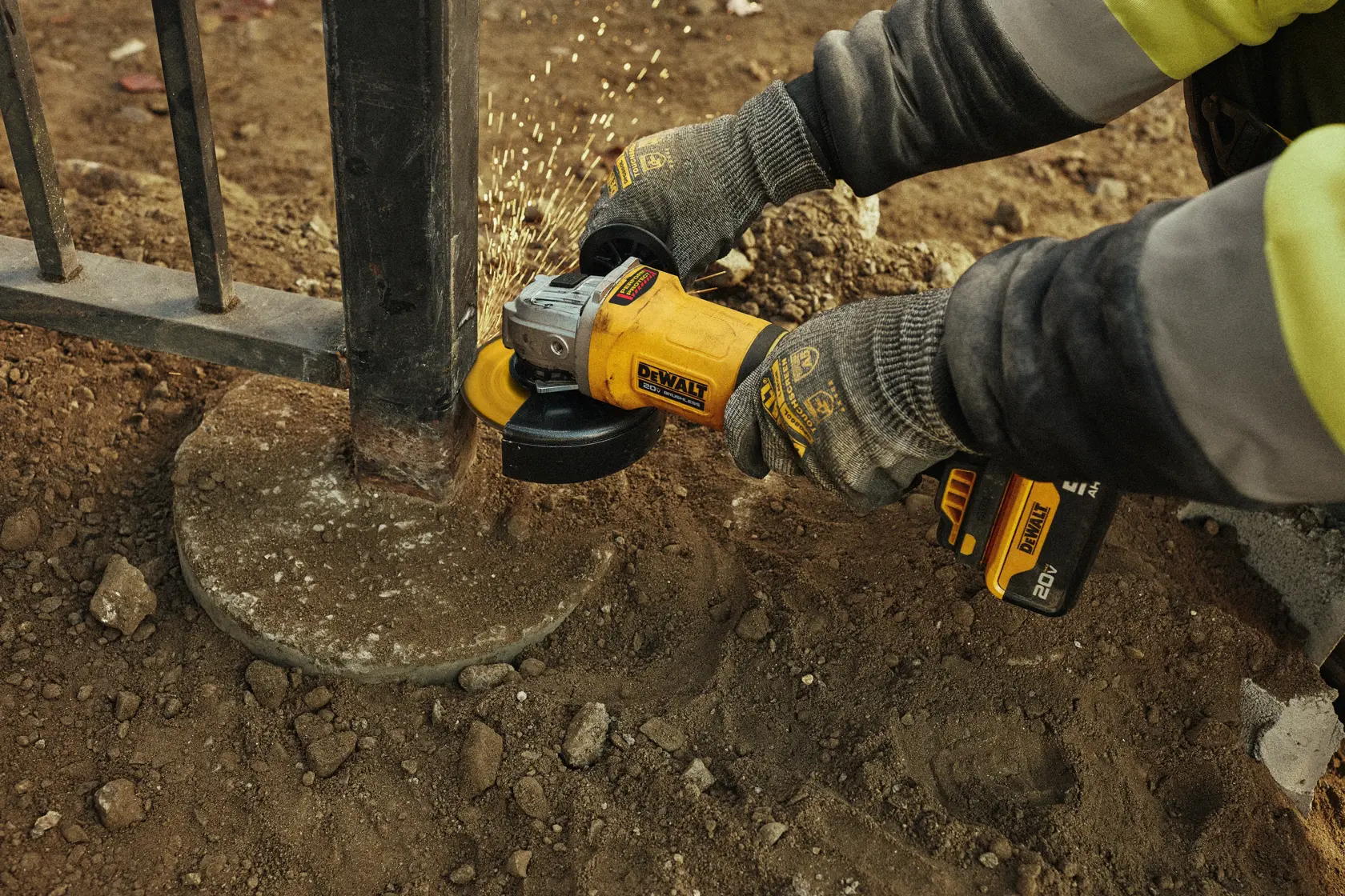 Person wearing gloves using a yellow DEWALT cordless angle grinder to grind metal on an outdoor fence post, with sparks flying.