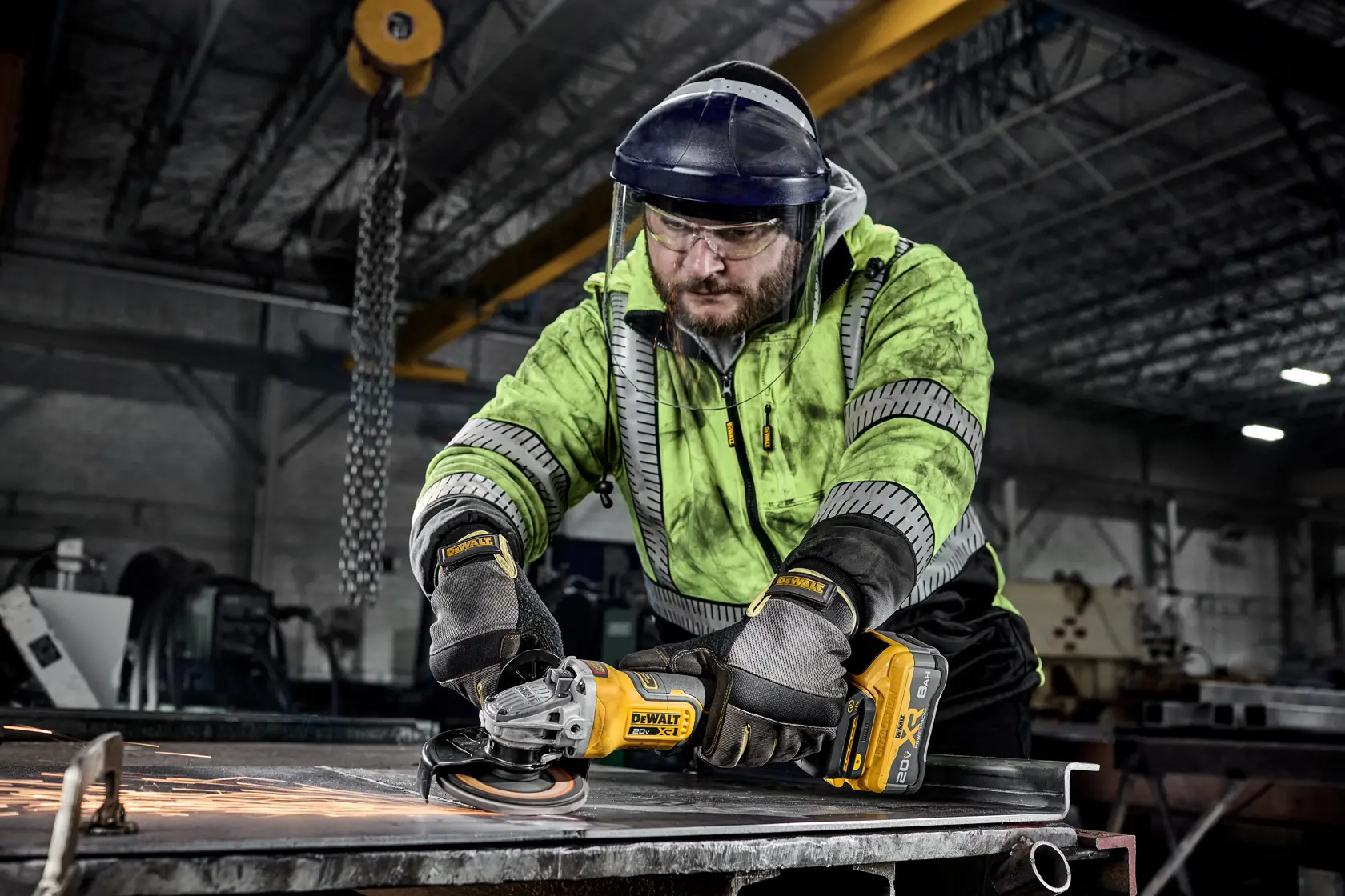 A factory worker using a DEWALT DCG410B cordless grinder to grind metal, wearing safety gloves, a high-visibility jacket, and a face shield. Sparks are visible from the grinding process. Industrial equipment and structural beams are in the background.