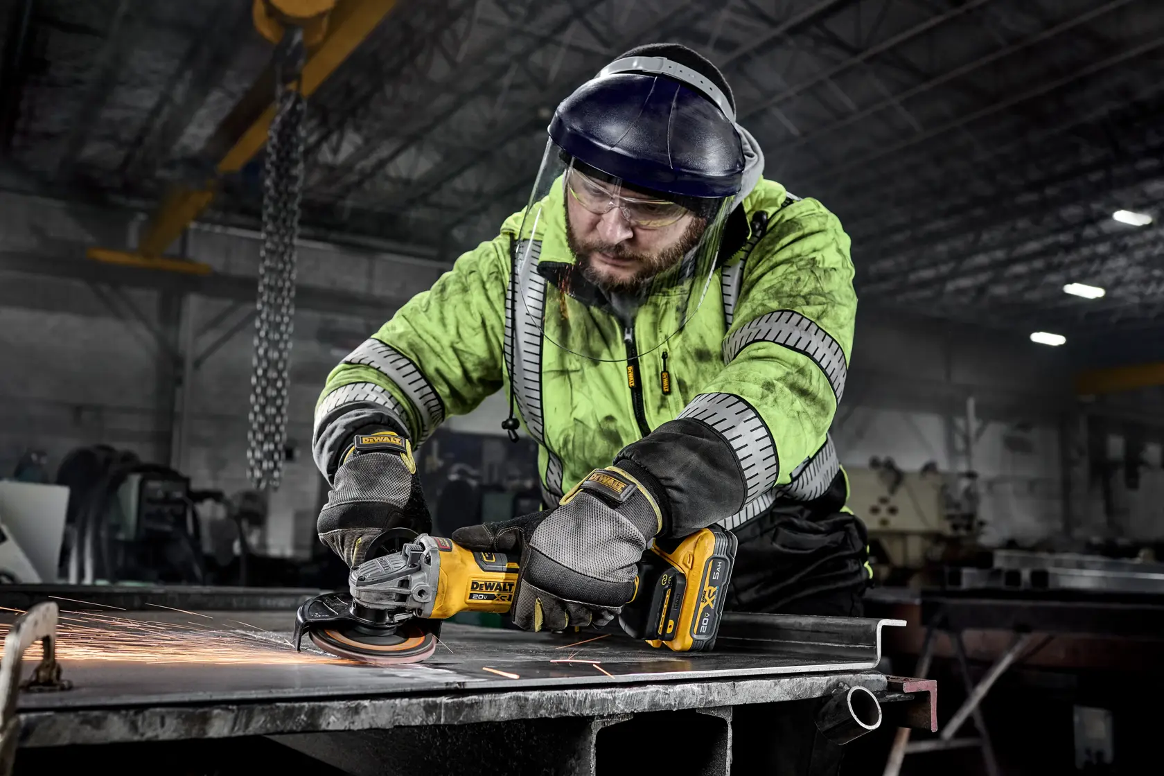 A worker in a safety jacket, gloves, and helmet is using a DEWALT cordless angle grinder to cut or grind metal in an industrial workshop. Sparks are visible as the tool is operating.