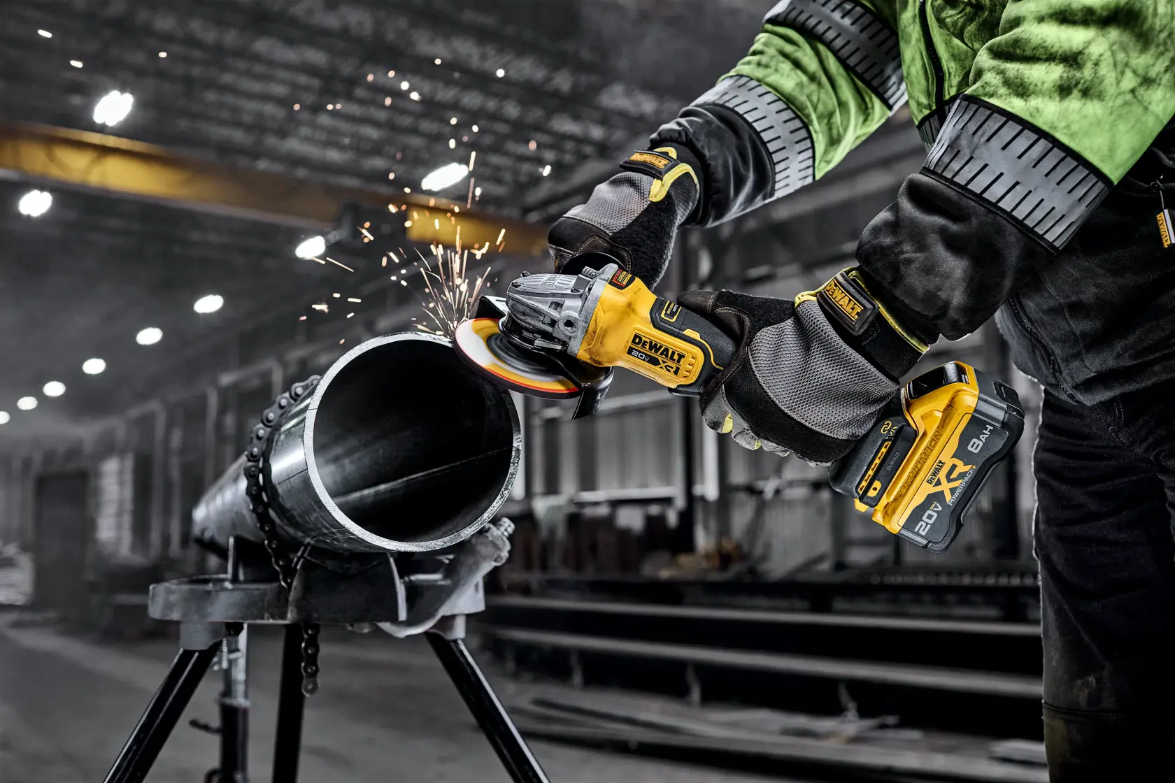 A DEWALT cordless angle grinder being used to cut a metal pipe, with sparks flying. The person operating the tool is wearing protective gloves and a green and black work jacket, in an industrial workshop setting.