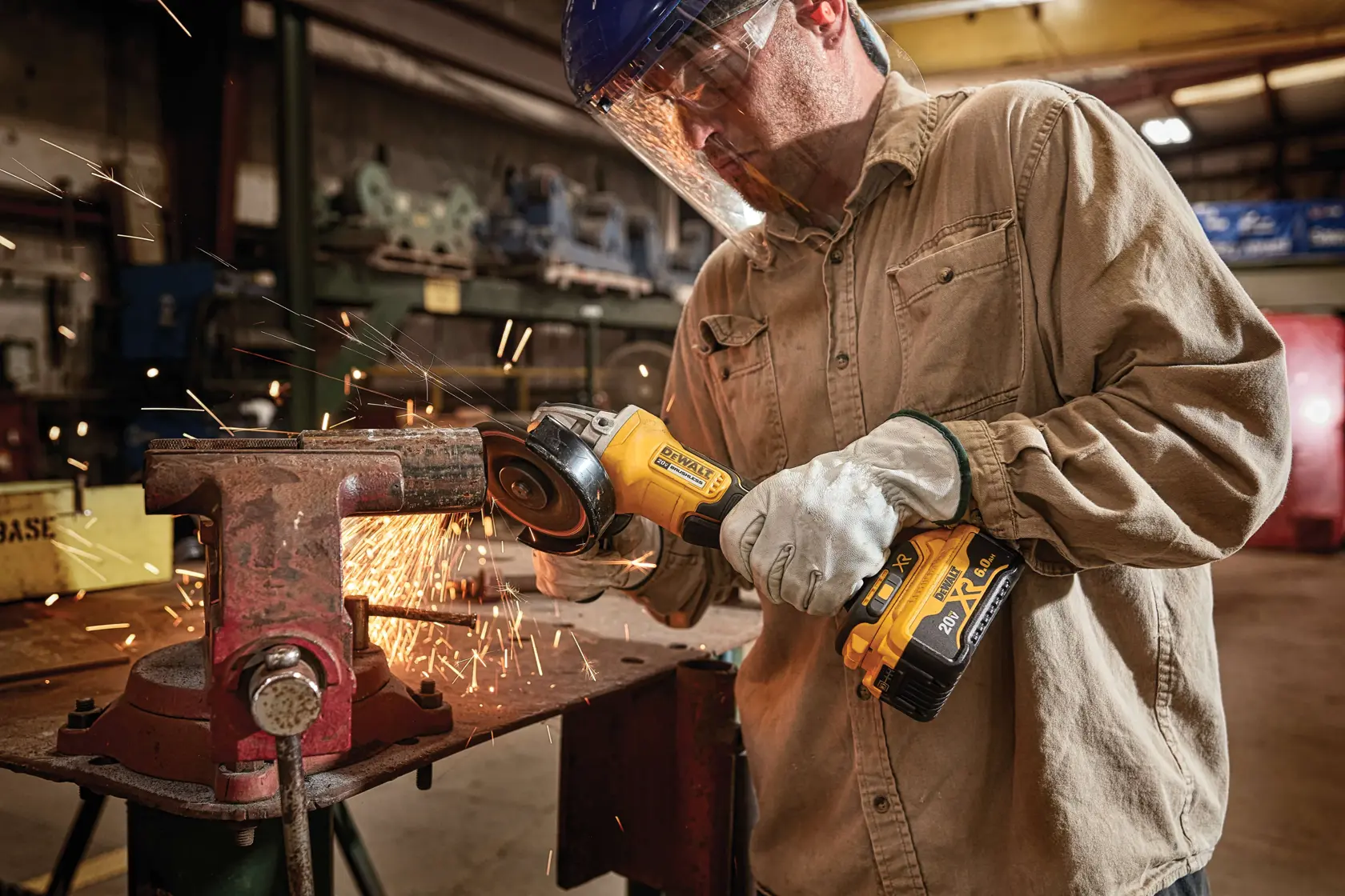 A person wearing protective gloves and a face shield is using a DEWALT DCG413R2 cordless angle grinder to cut or grind a metal pipe held in a vise. Sparks are flying from the grinding process, and the setting appears to be an industrial workshop.